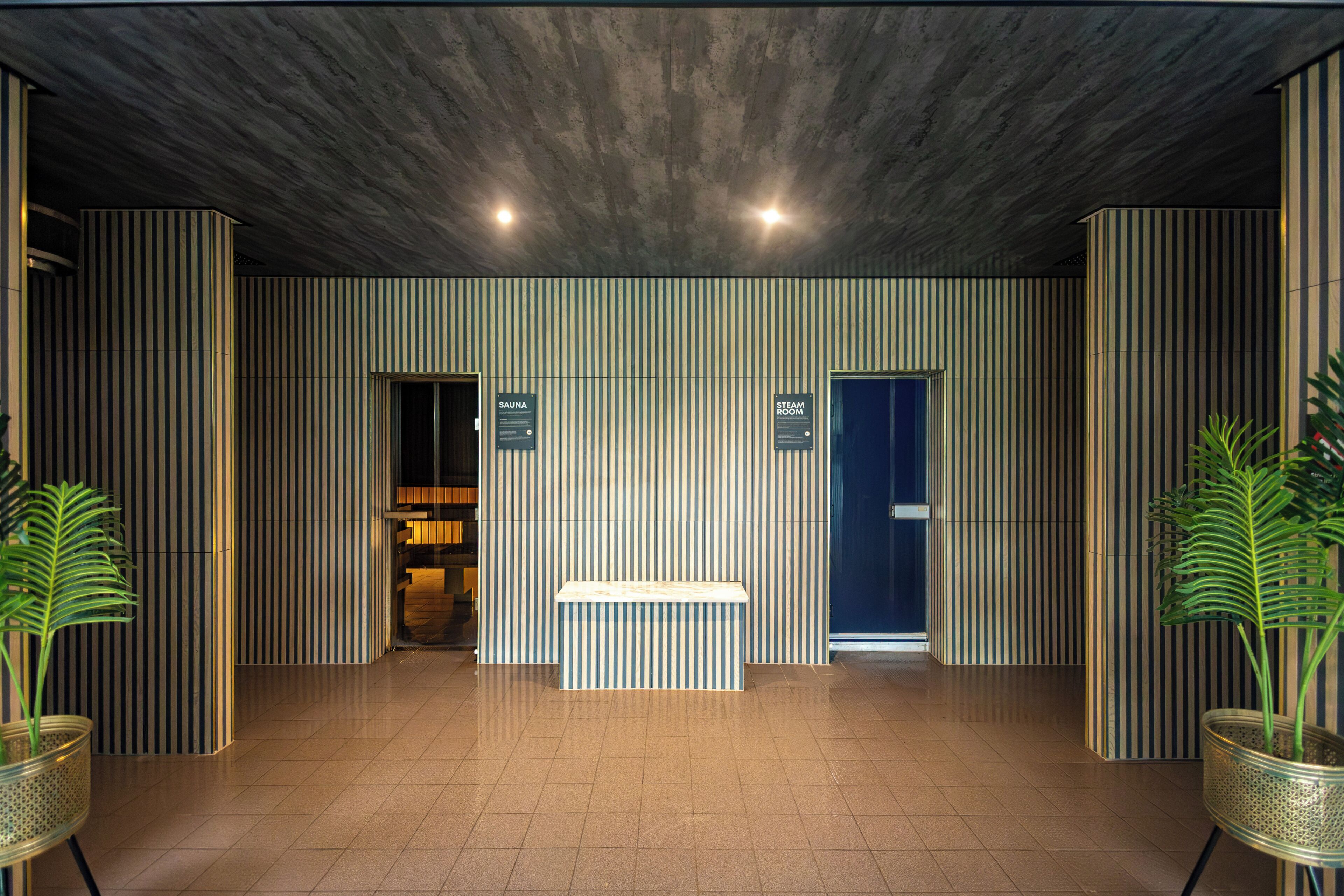 A reception area with a bench, two doors labeled 'SAUNA' and 'STEAM ROOM,' and vertical striped walls.