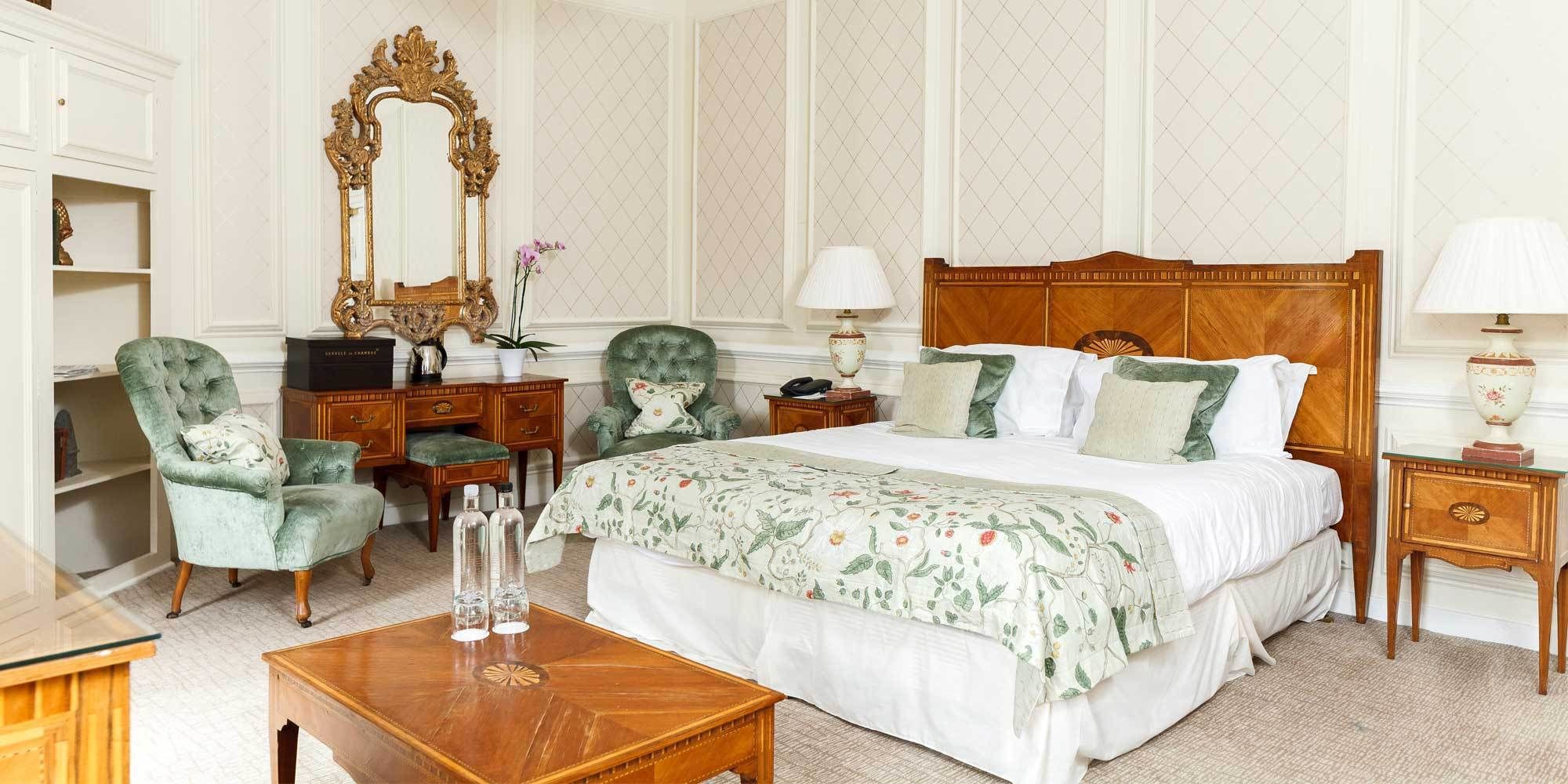 A hotel room with a large bed, two armchairs, a wooden desk, a mirror, and a nightstand with lamps.