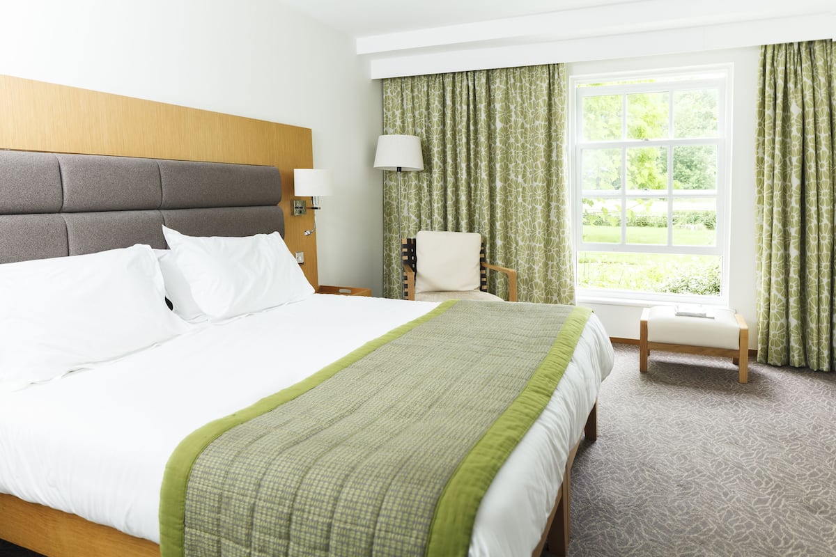 A hotel room with a large bed, a wooden headboard, a window with curtains, a chair, and a nightstand with lamps.