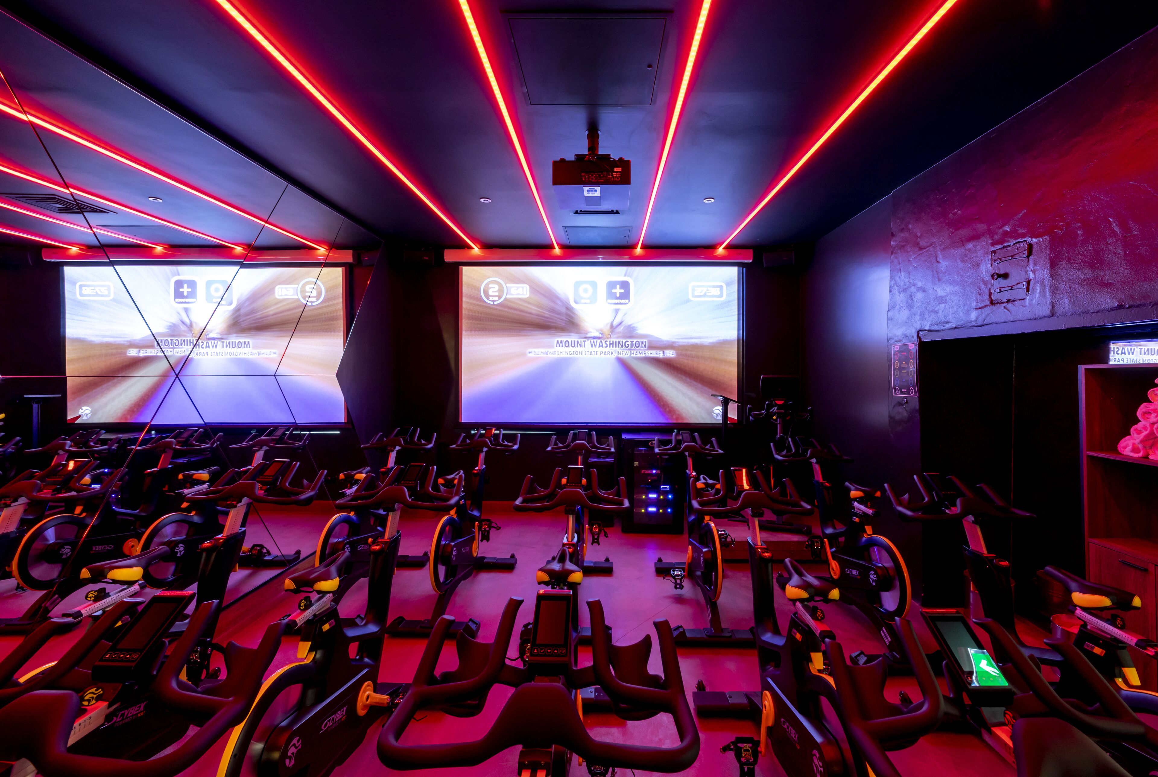 A gym with stationary bikes arranged in rows, a large screen displaying a bowling game, and red lighting.