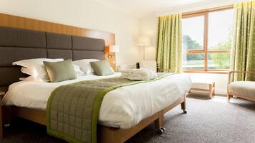 Une chambre d’hôtel avec un grand lit, une tête de lit en bois, une table de chevet avec une lampe, une chaise et une vue sur la verdure par la fenêtre.