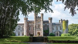 A historic stone building with multiple towers and a central entrance, surrounded by well-maintained lawns and trees.