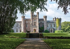 A historic stone building with multiple towers and a central entrance, surrounded by well-maintained lawns and trees.