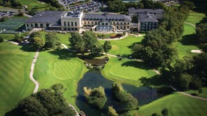 An aerial view of a golf course with a clubhouse, a pond, and surrounding greenery.