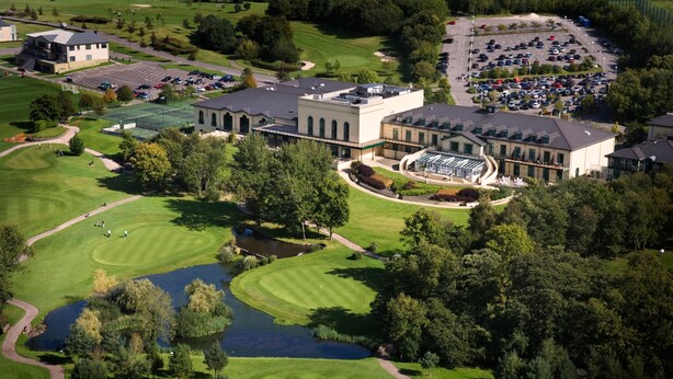 An aerial view of a golf course with a clubhouse, parking lot, and surrounding greenery.