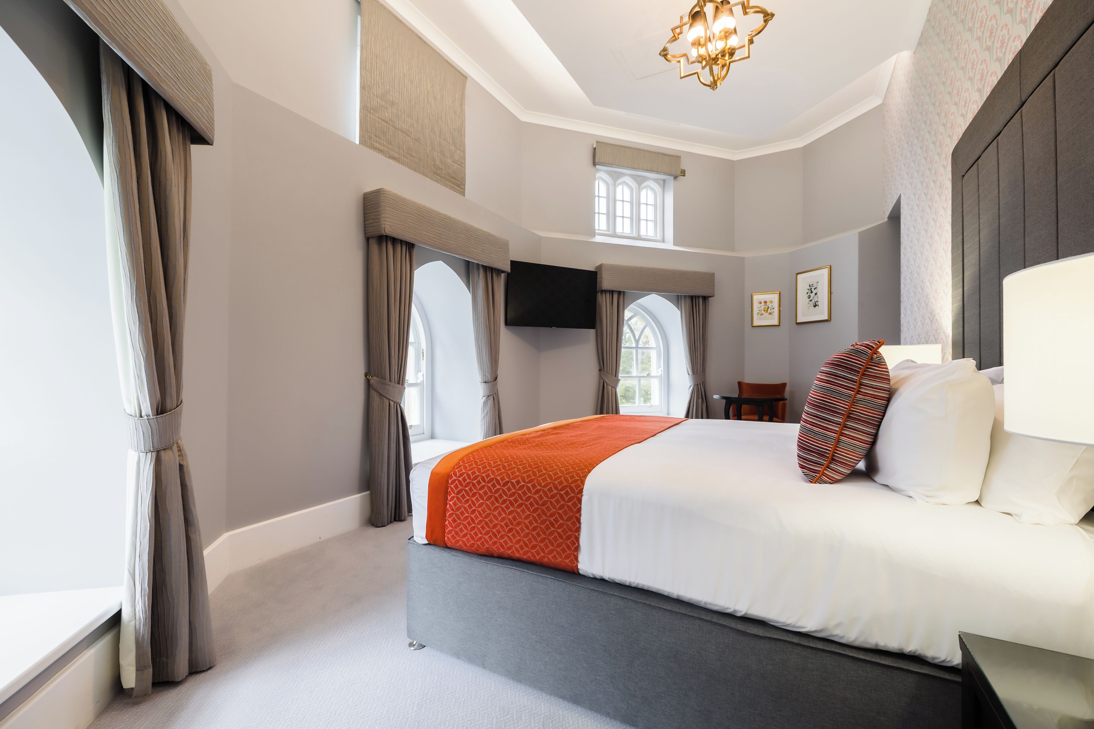 A modern hotel room with a large bed, a flat-screen TV, a window with curtains, and a chandelier.