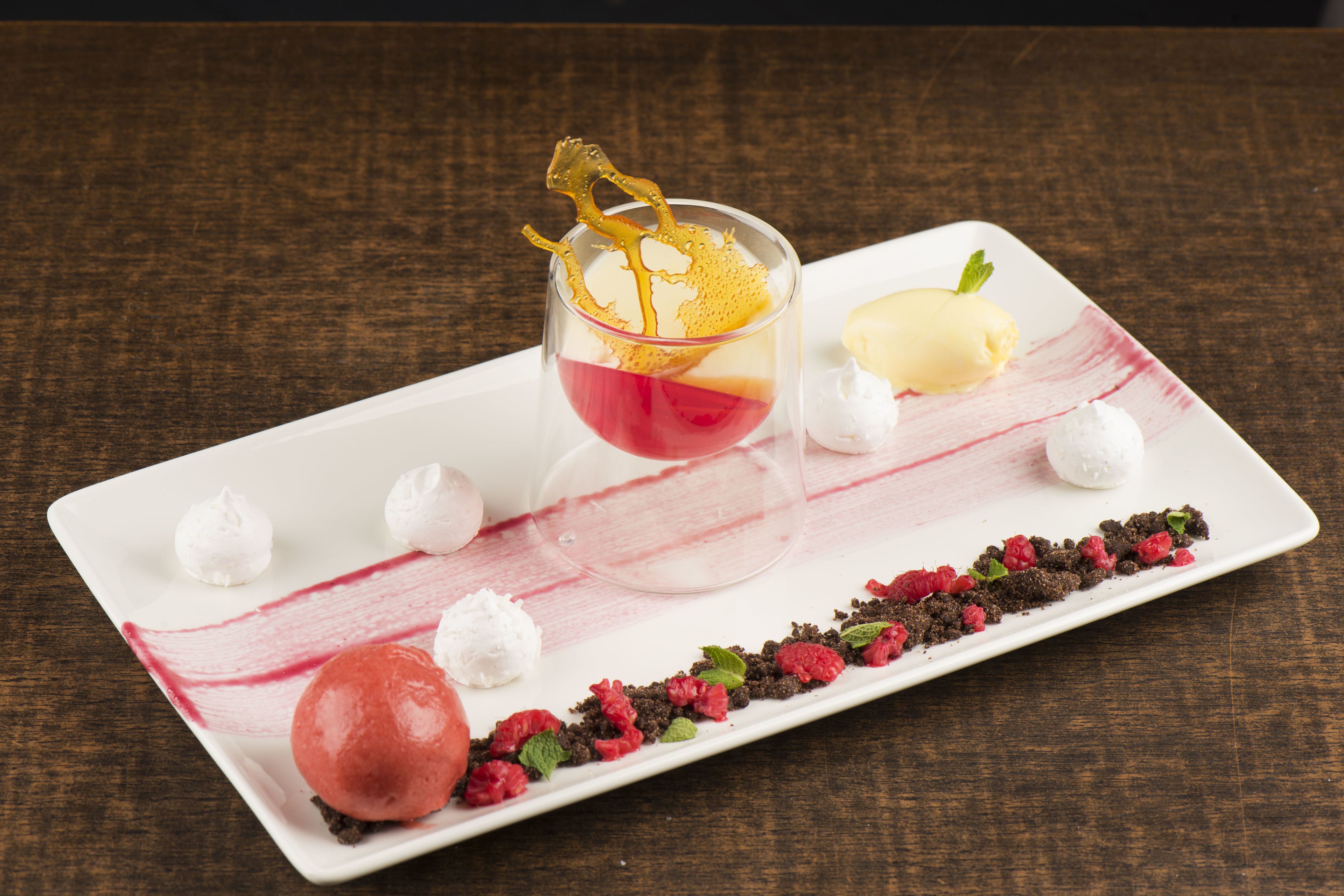Un plato de postre con varios elementos, incluyendo un vaso con líquido rojo, una bola de helado y adornos como frambuesas y hojas de menta.