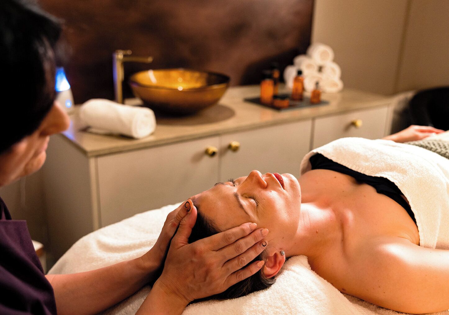A person receiving a facial treatment in a spa setting with a massage table, a bowl, and various bottles on a counter.