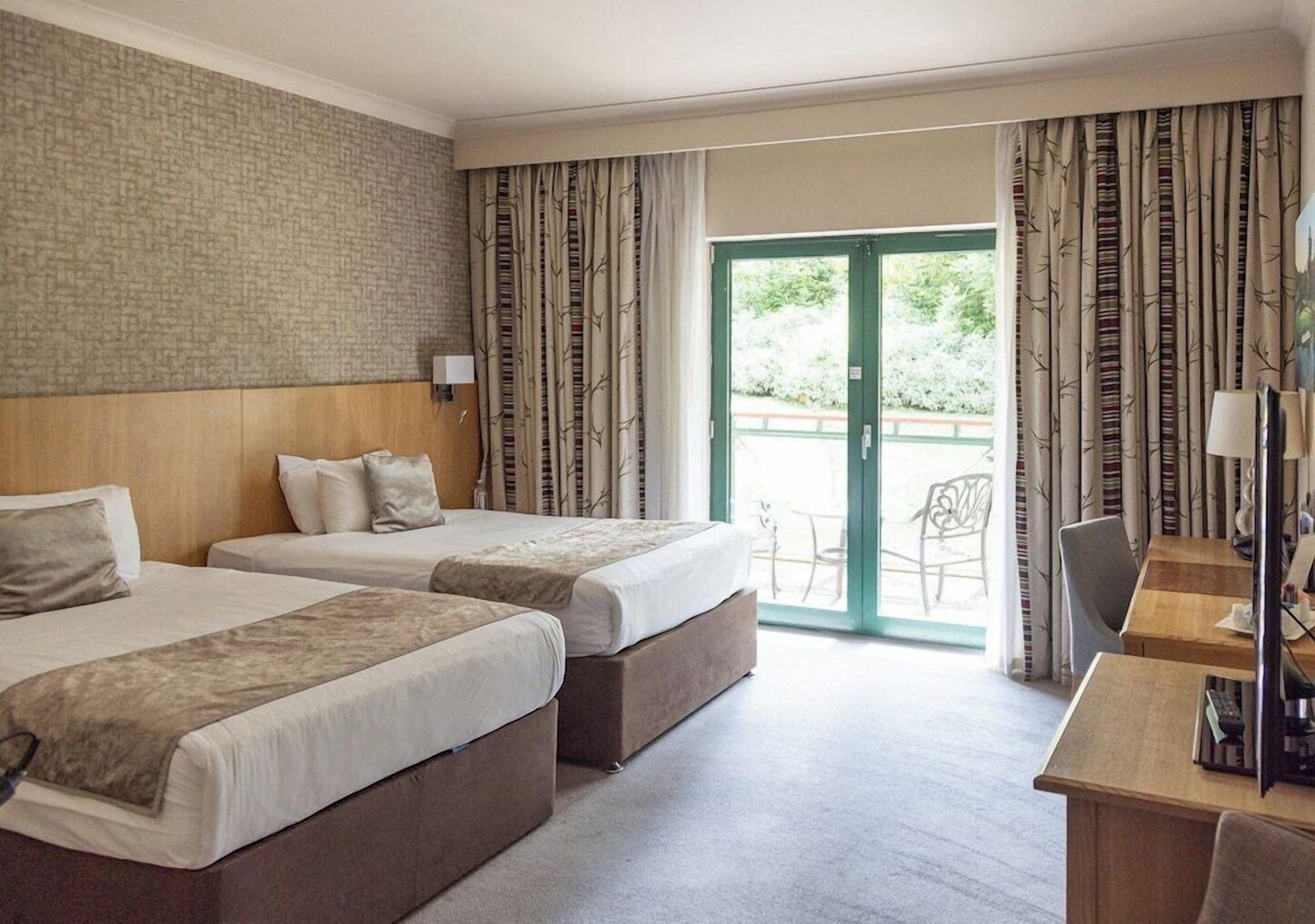 Quarto de hotel com duas camas, cabeceira de madeira, uma escrivaninha com computador e vista para o exterior através de uma porta de vidro.