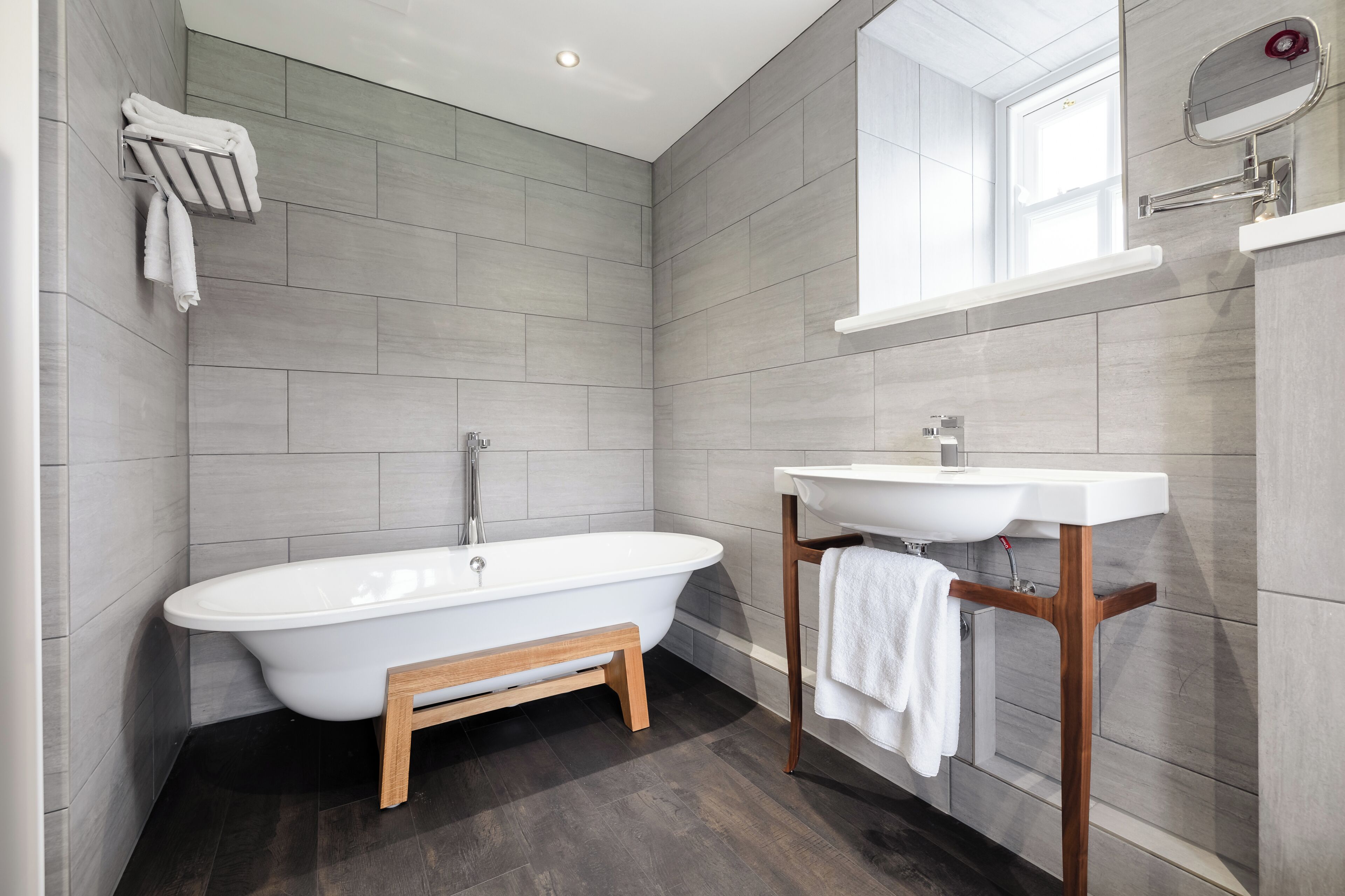 Un baño moderno con una bañera independiente, un banco de madera, un lavabo blanco y un espejo sobre él.