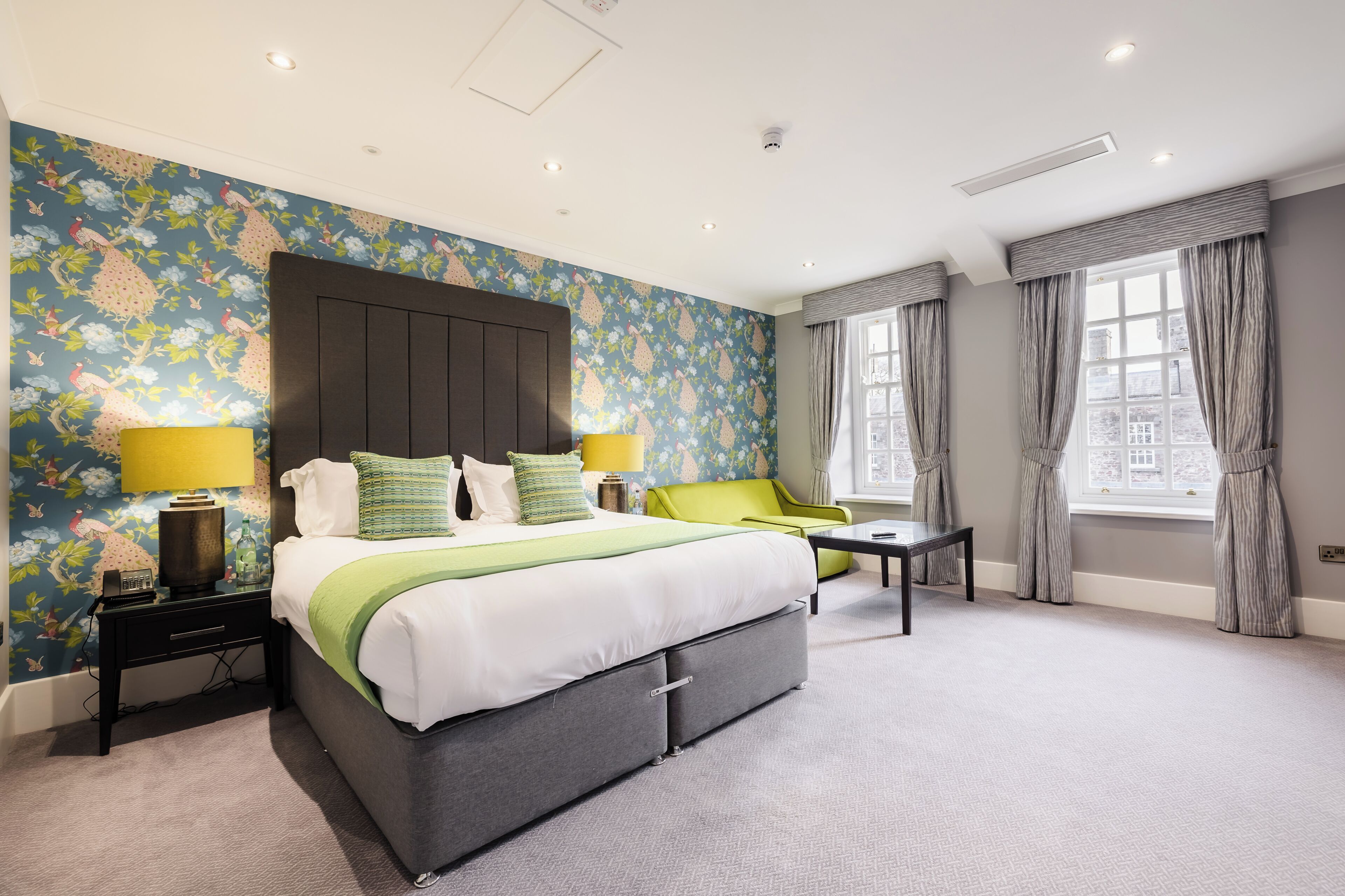 A hotel room with a large bed, a bedside table, a chair, a window with curtains, and a floral wallpaper.