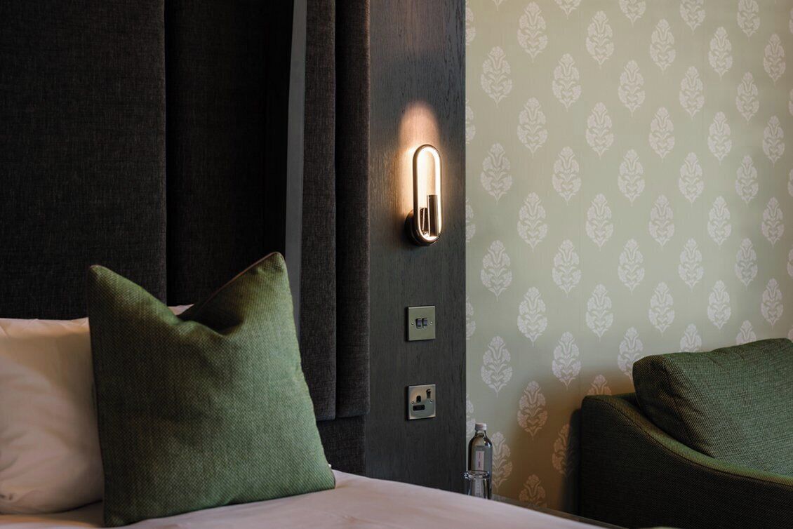 A hotel room with a bed, green pillows, a wall-mounted light, and a patterned wallpaper.