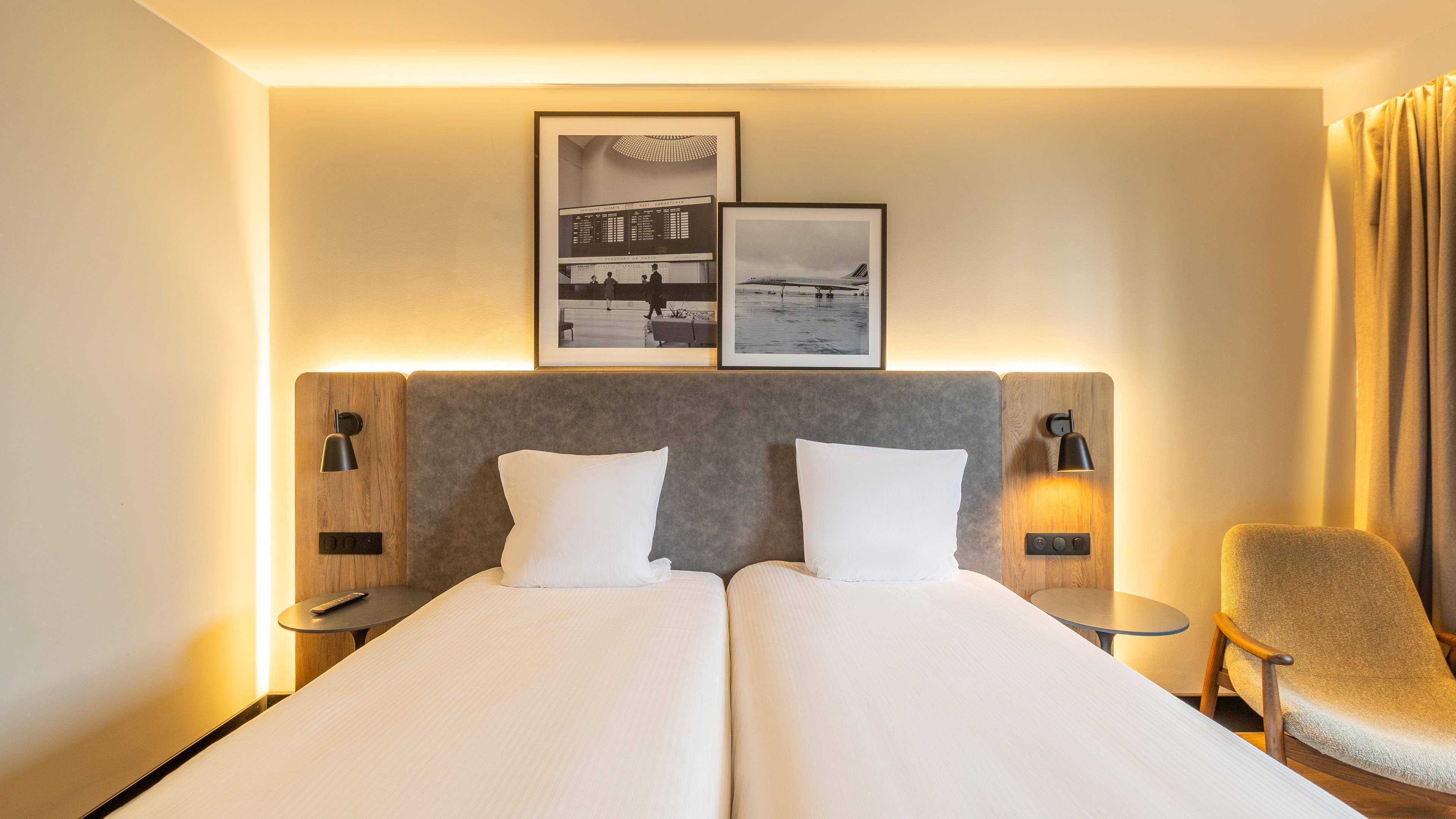 A hotel room with two beds, a chair, and two wall-mounted lamps.