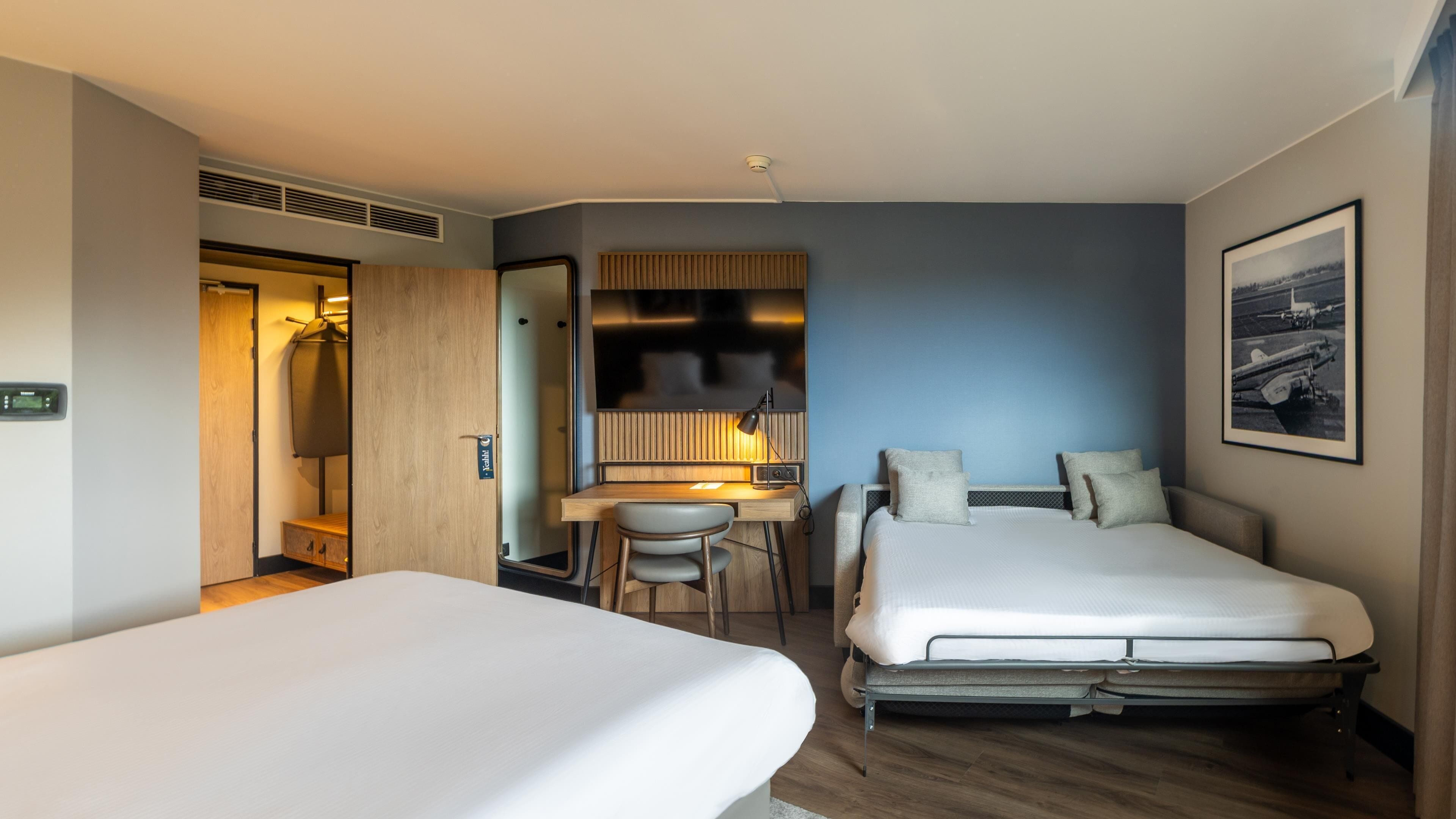 A hotel room with two beds, a desk, a chair, a wardrobe, a television, and a framed picture on the wall.