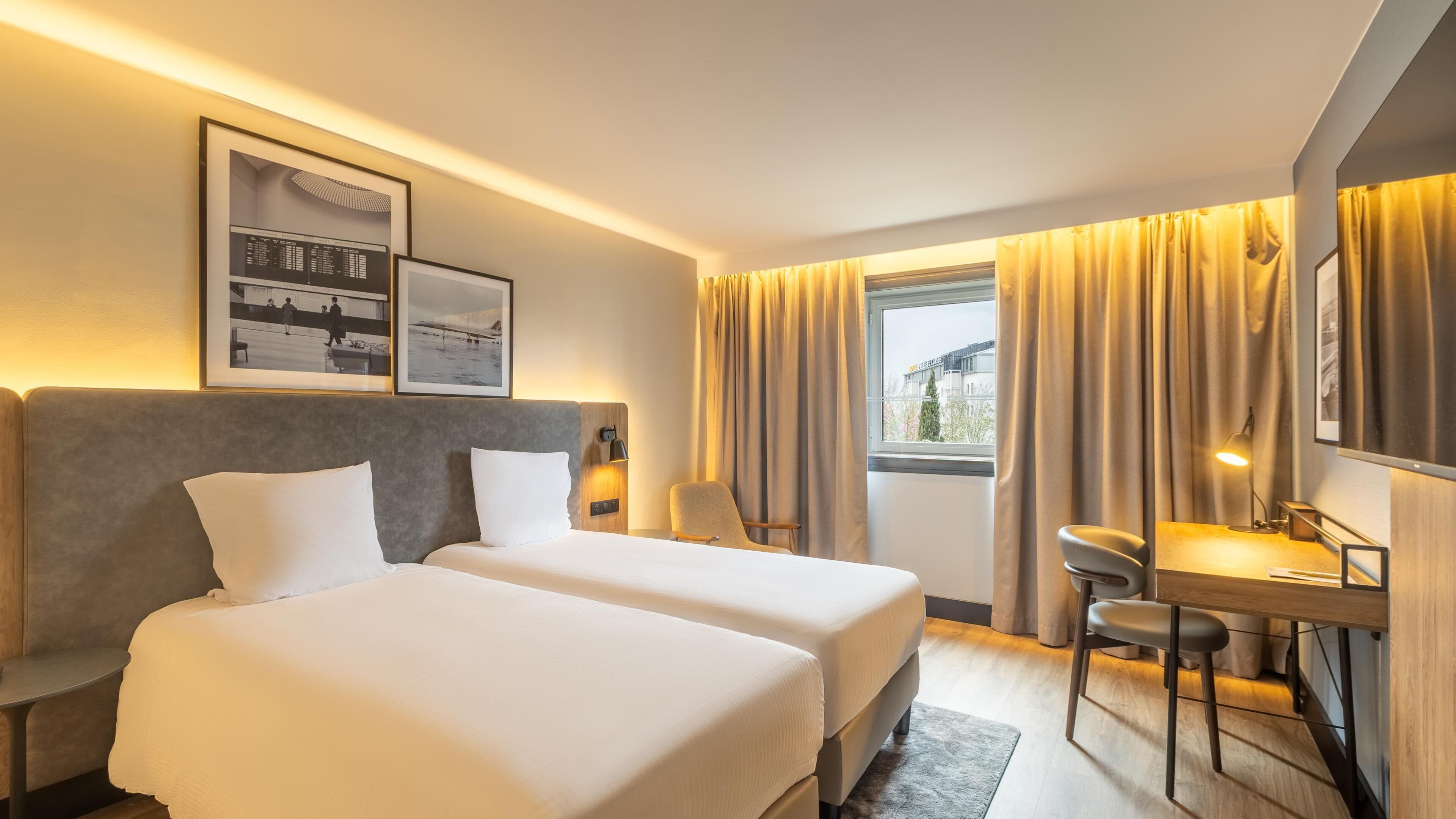A hotel room with two beds, a desk, a chair, a lamp, and a window with curtains.