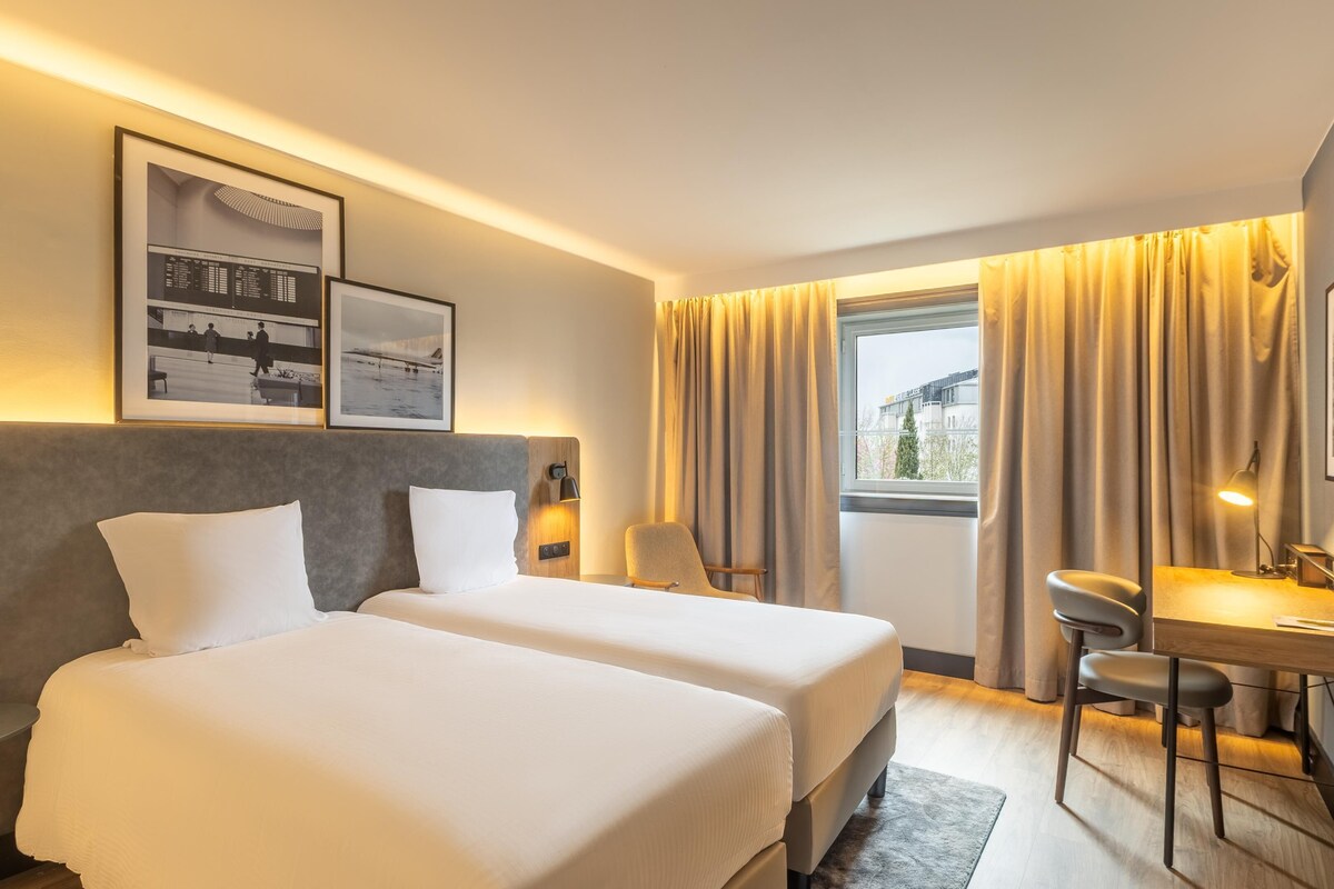 Une chambre d’hôtel avec deux lits, un bureau, une chaise, une lampe et une fenêtre avec des rideaux.