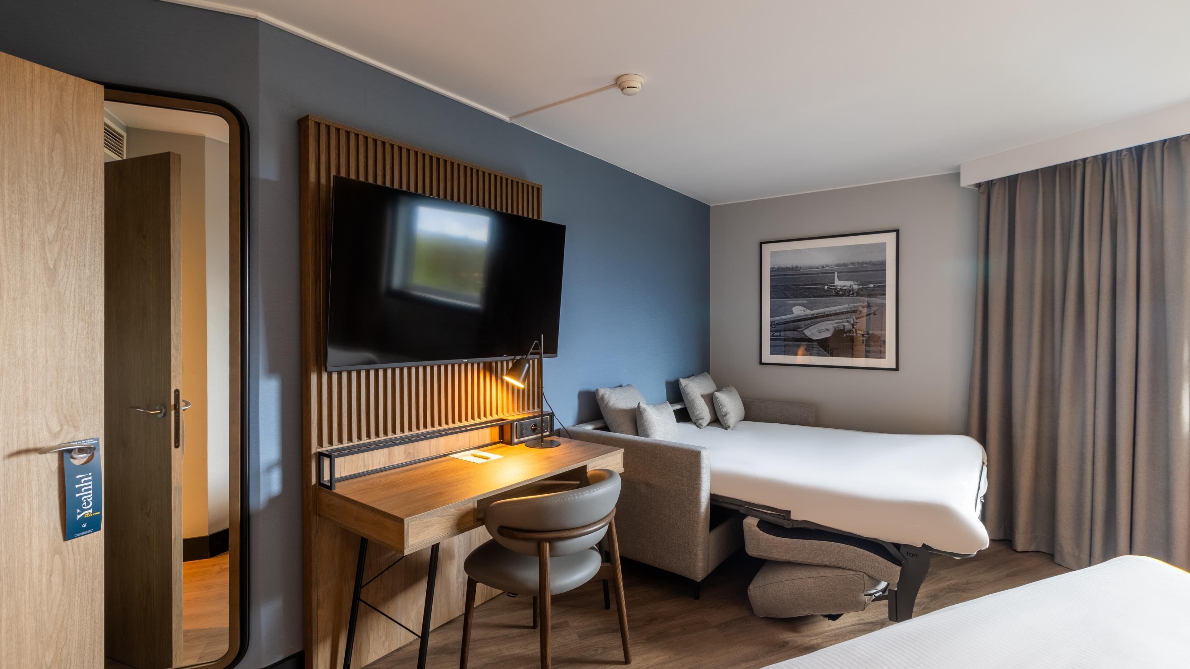 Una habitación de hotel con una cama, un escritorio con una silla, un televisor y un cuadro en la pared.