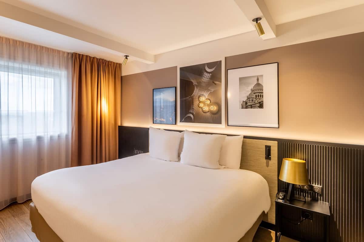 Et hotelværelse med en stor seng, et natbord med en lampe og billeder i rammer på væggen.
