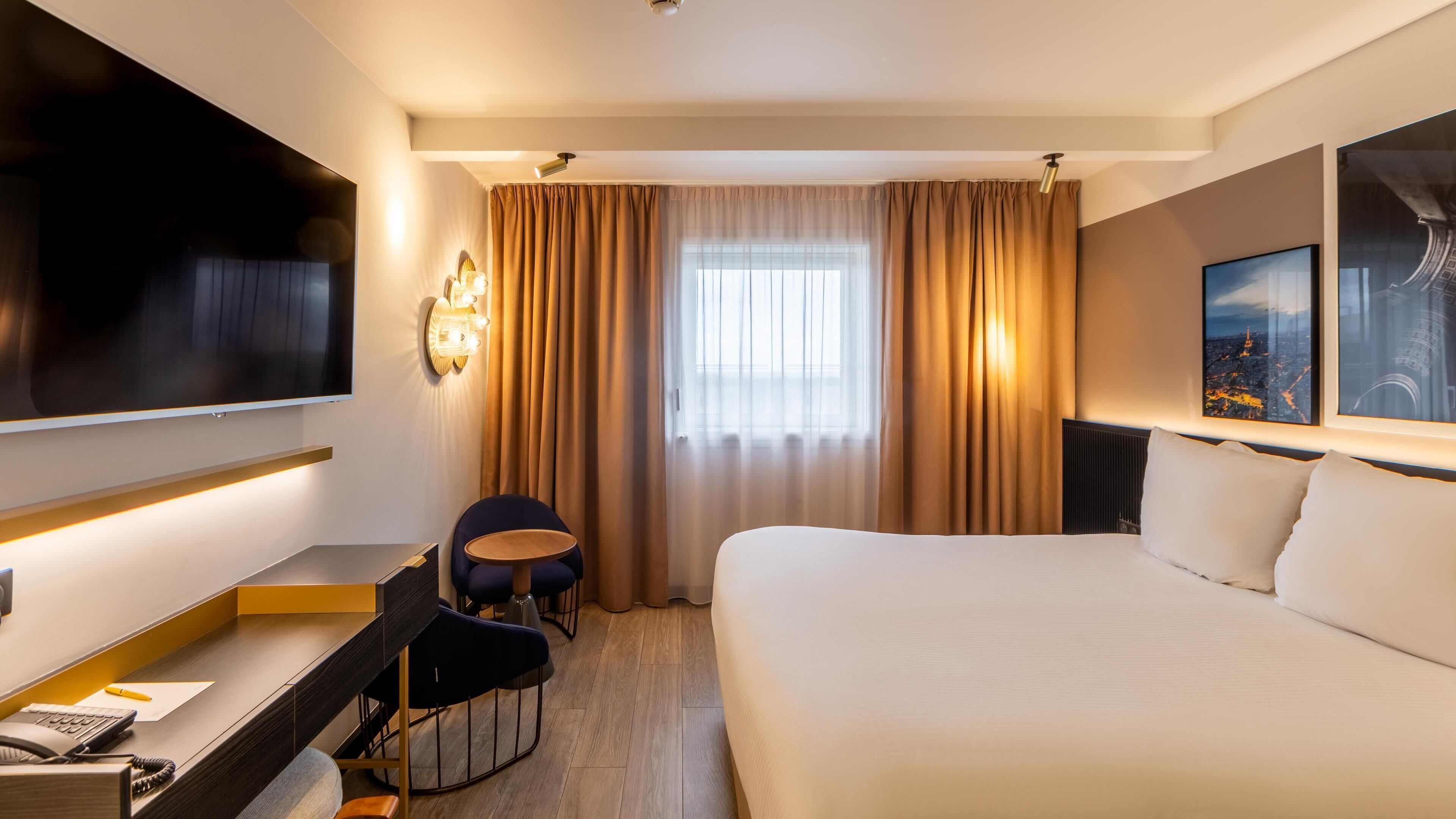 A hotel room with a bed, desk, chair, TV, curtains, and a cityscape view.