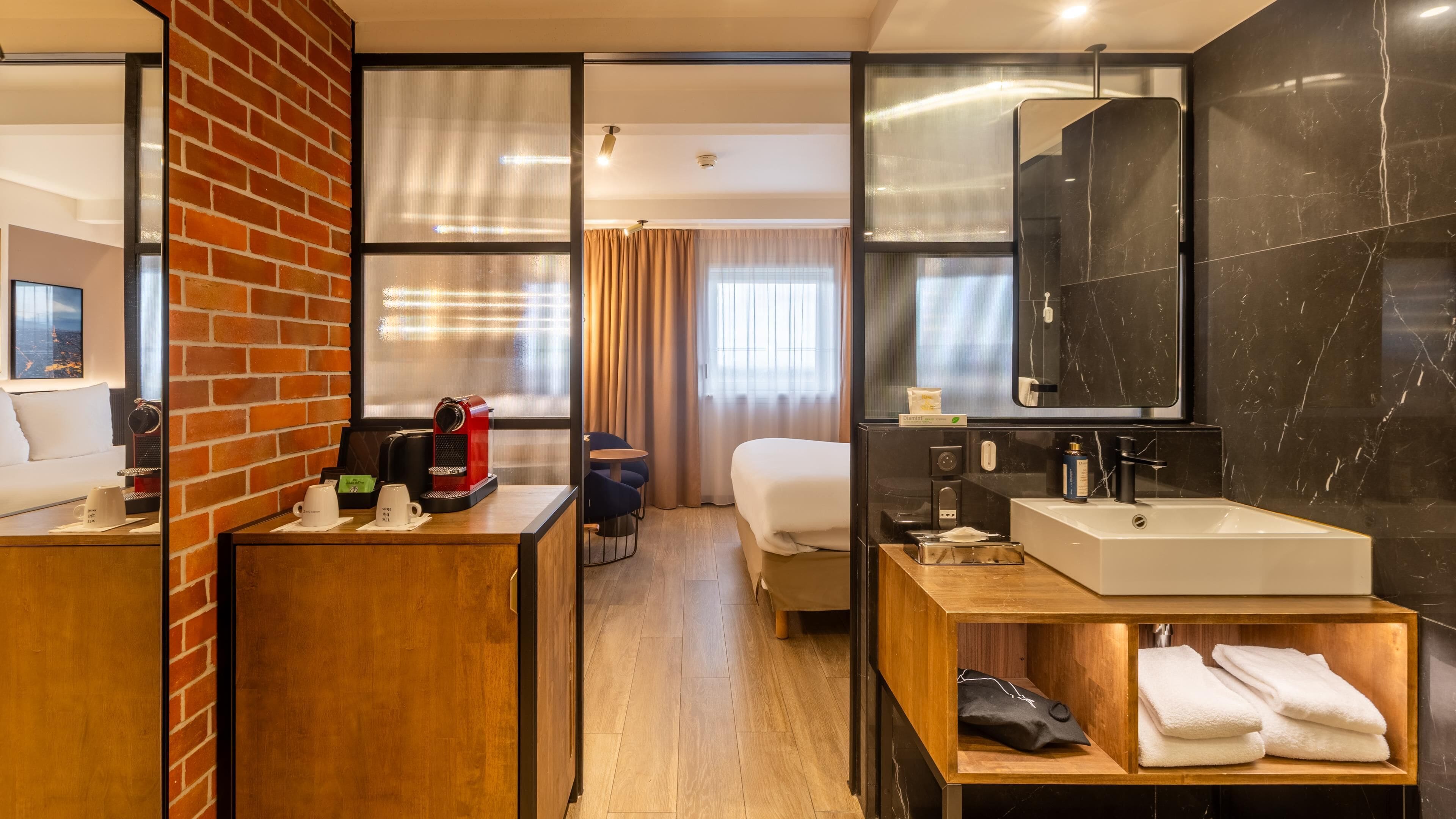 Une chambre d’hôtel avec un lit, un bureau en bois avec une machine à café, une salle de bain avec un grand miroir et un plancher en bois.