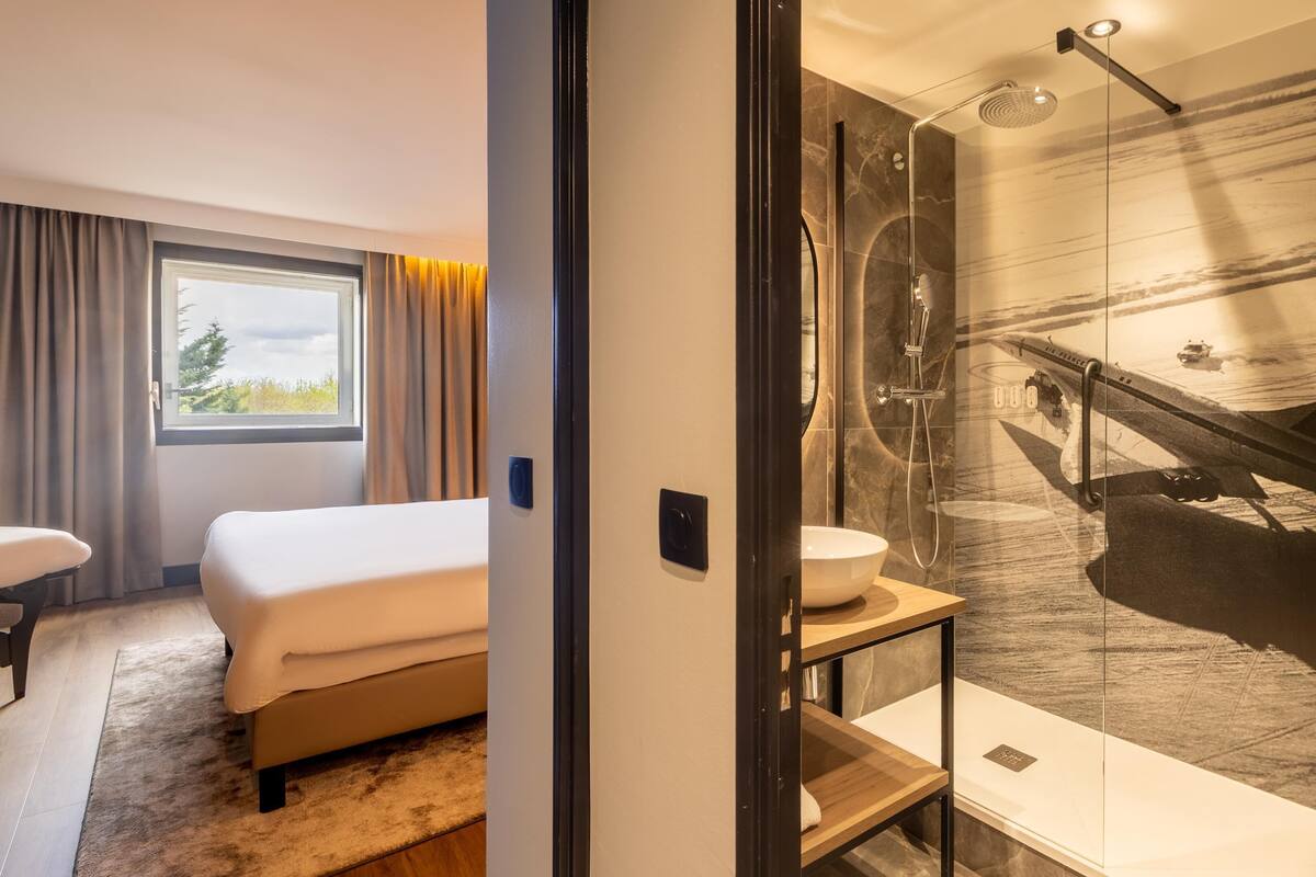 Et hotelværelse med en seng, et badeværelse med et stort spejl, en vask og en bruser.