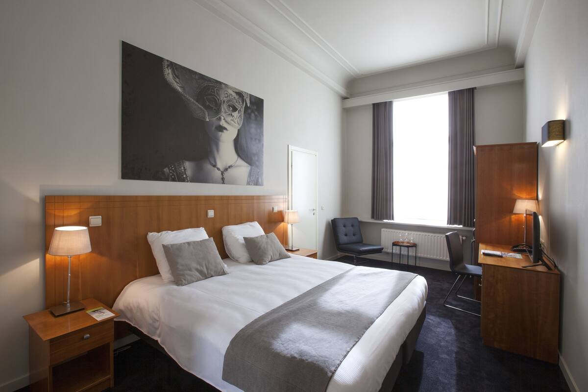Una habitación de hotel con una cama grande, lámparas de noche, un escritorio, una silla y un cuadro grande en la pared.