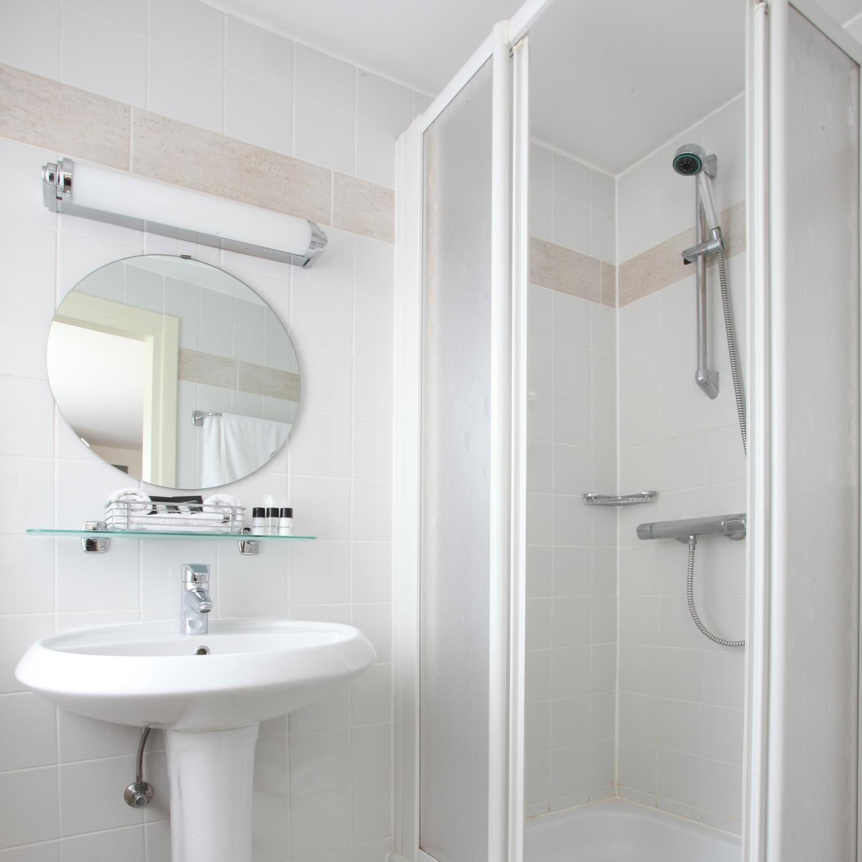 A modern bathroom with a white sink, a glass shelf with toiletries, a round mirror, and a walk-in shower with a handheld showerhead.