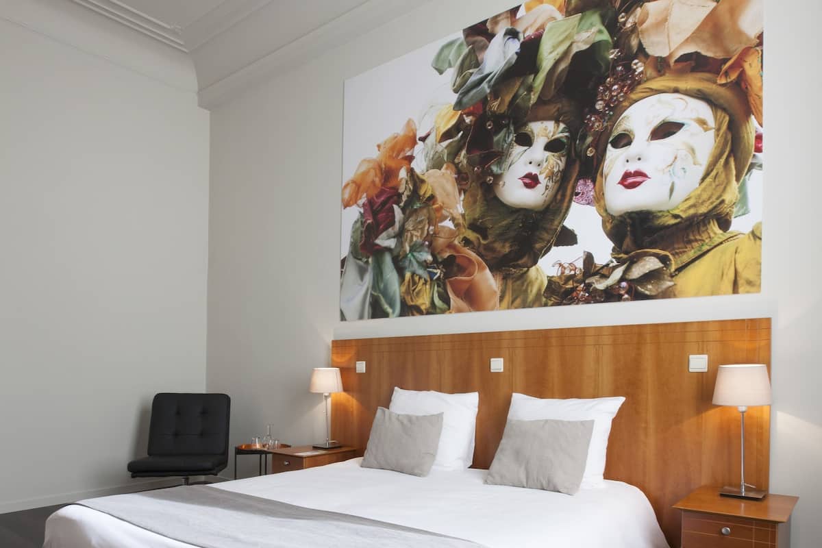 Una habitación de hotel moderna con una cama grande, cabecera de madera, dos mesitas de noche con lámparas y un cuadro abstracto de gran tamaño en la pared.
