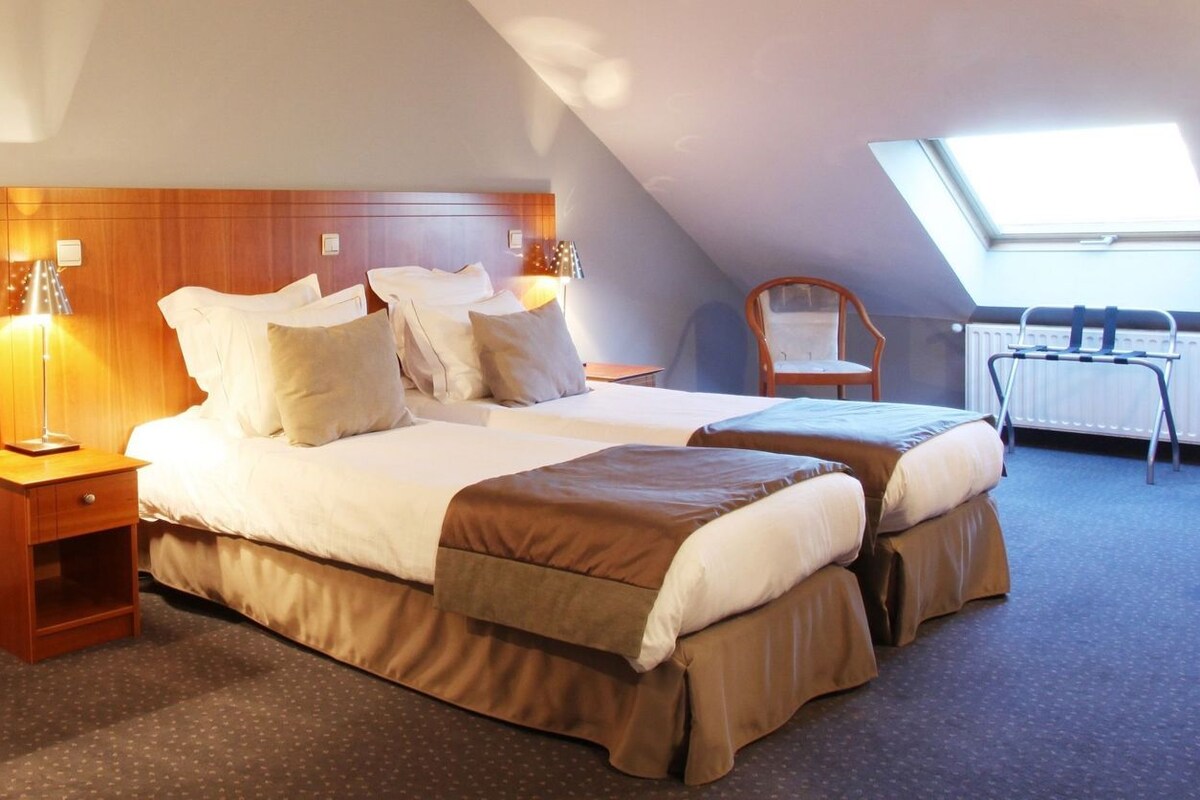 Une chambre d’hôtel avec deux lits, une tête de lit en bois, un miroir, une petite table, une chaise et une lucarne.