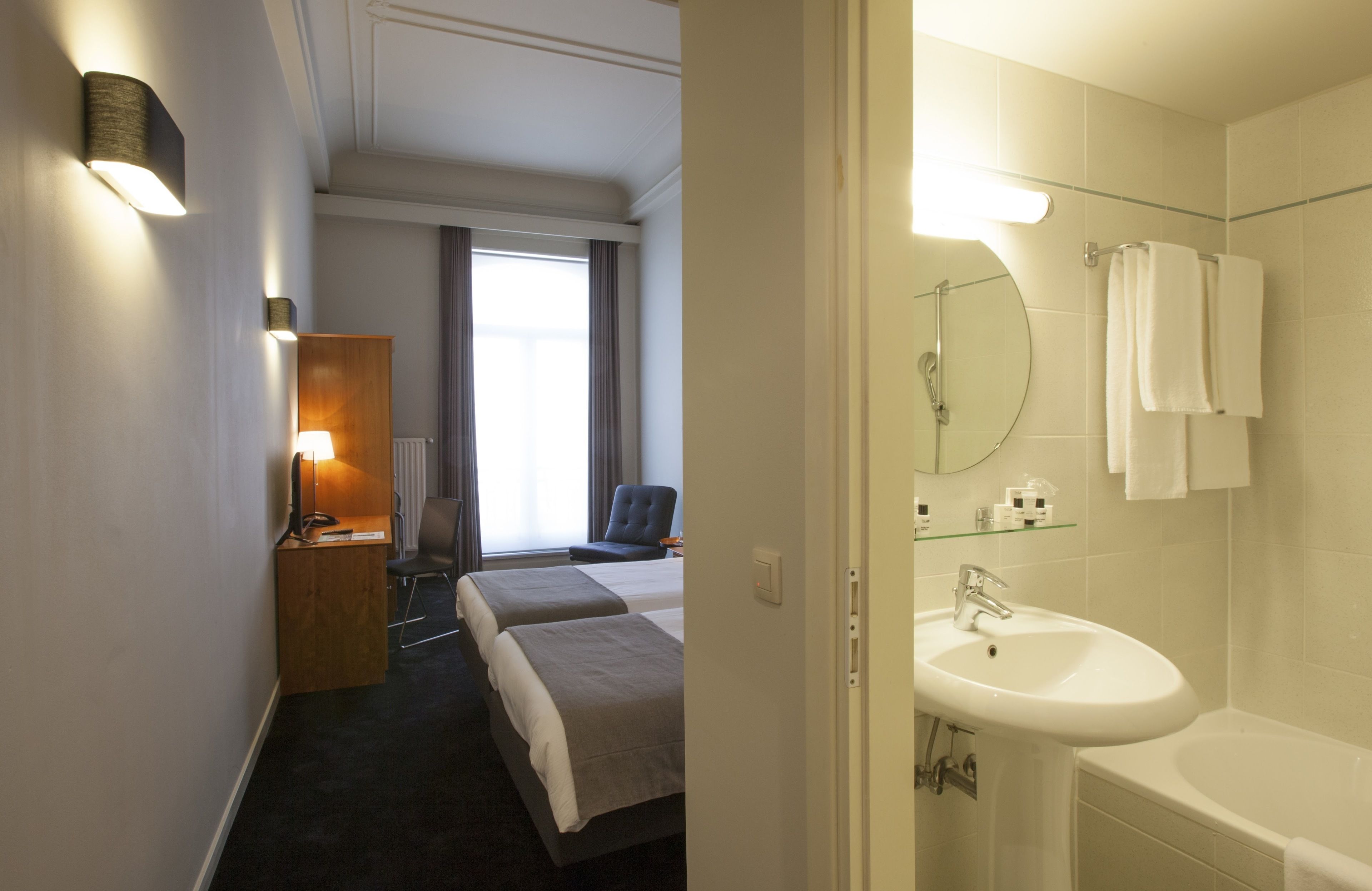 Habitación de hotel con cama, mesita de noche, lámpara, silla y vistas al exterior.