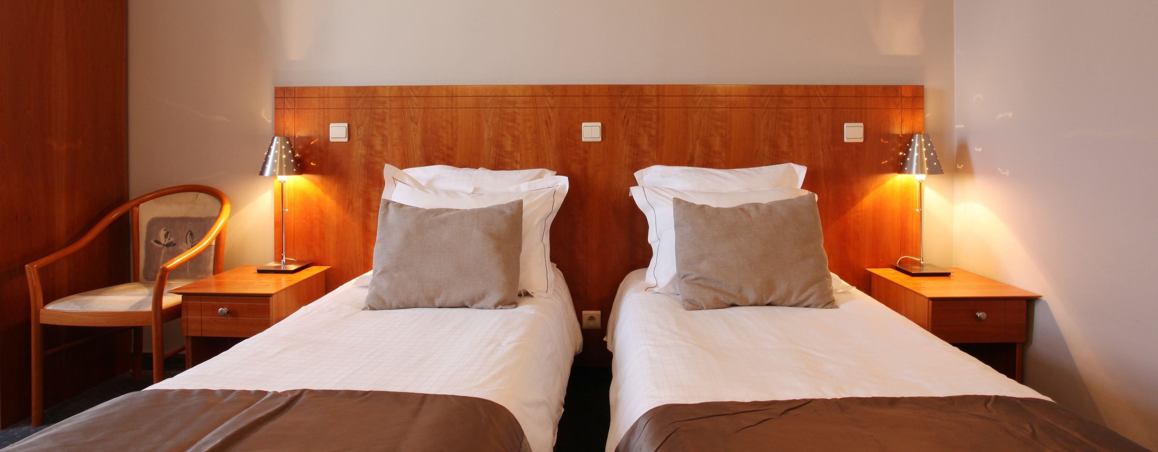 Habitación de hotel con dos camas individuales, cabecero de madera, lámparas de noche, una silla de madera y una mesita.