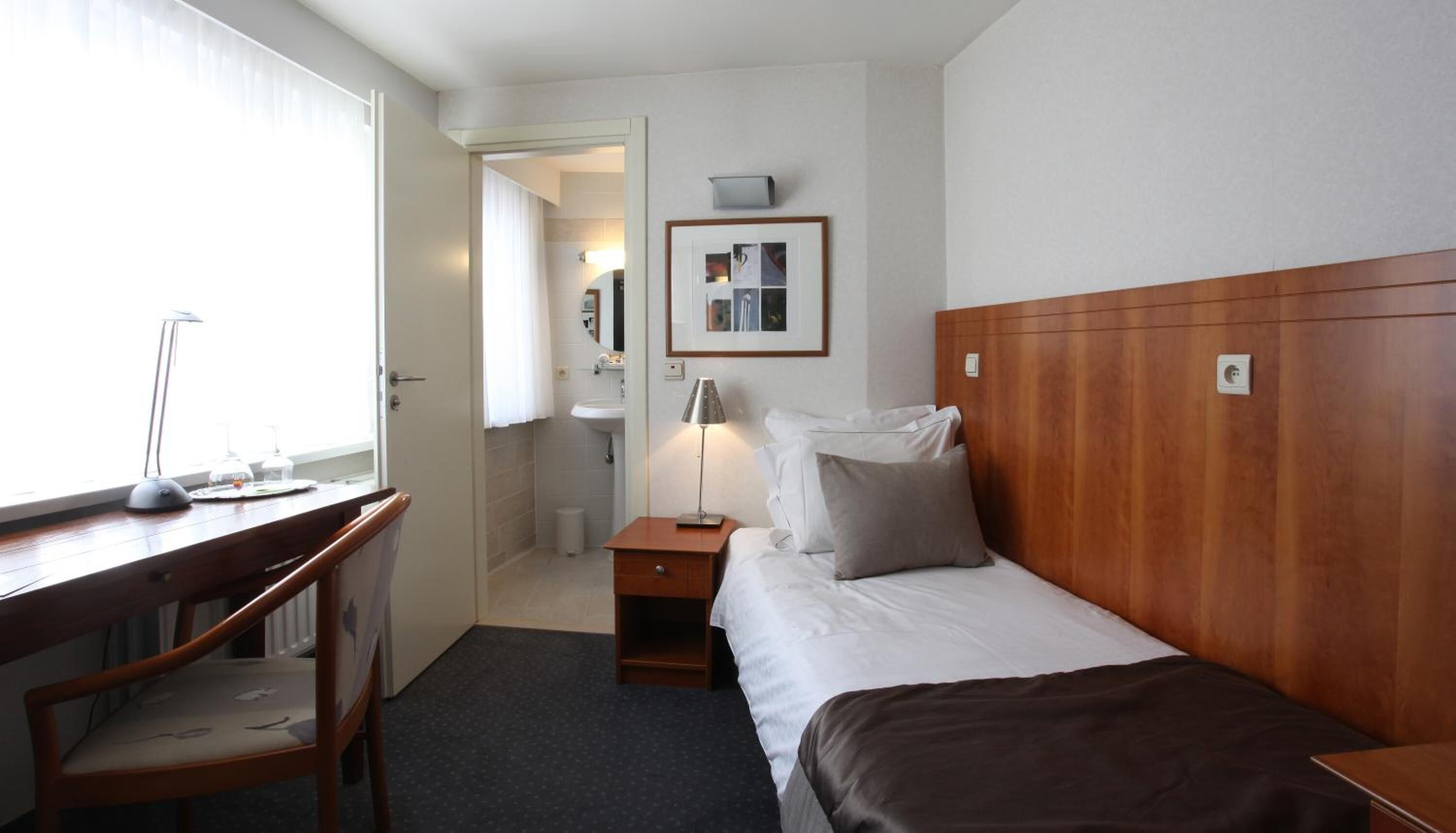A hotel room with a wooden headboard, a desk with a chair, a bed with pillows, a nightstand, and a framed picture on the wall.