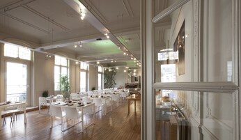 A spacious hotel dining area with multiple tables and chairs, large windows, and a mirror reflecting the room.