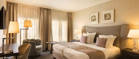 A hotel room with a large bed, two bedside tables with lamps, a desk with a chair, a round armchair, and a TV.