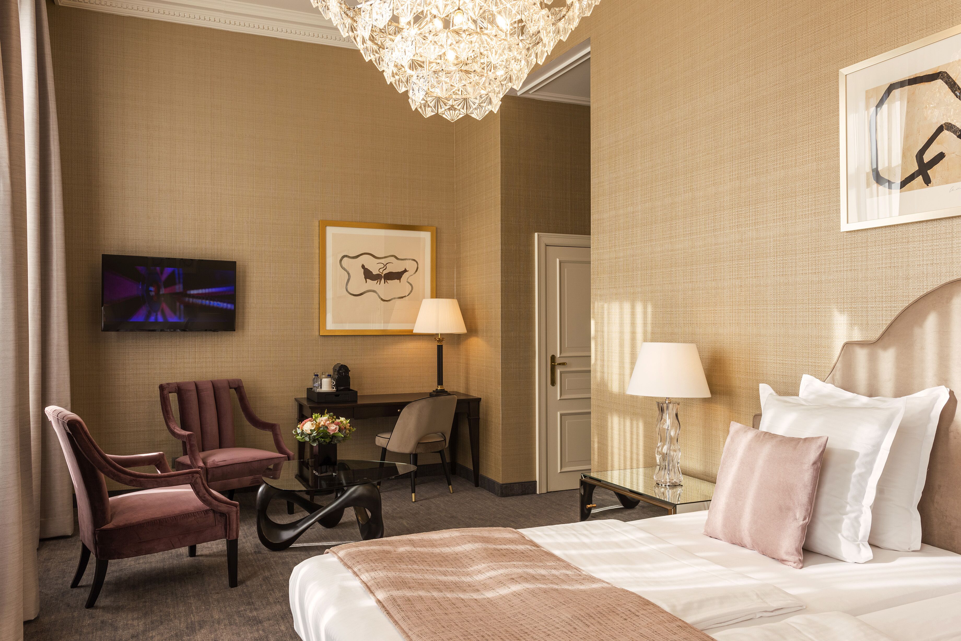 A hotel room with a bed, two chairs, a desk, a TV, and a chandelier.