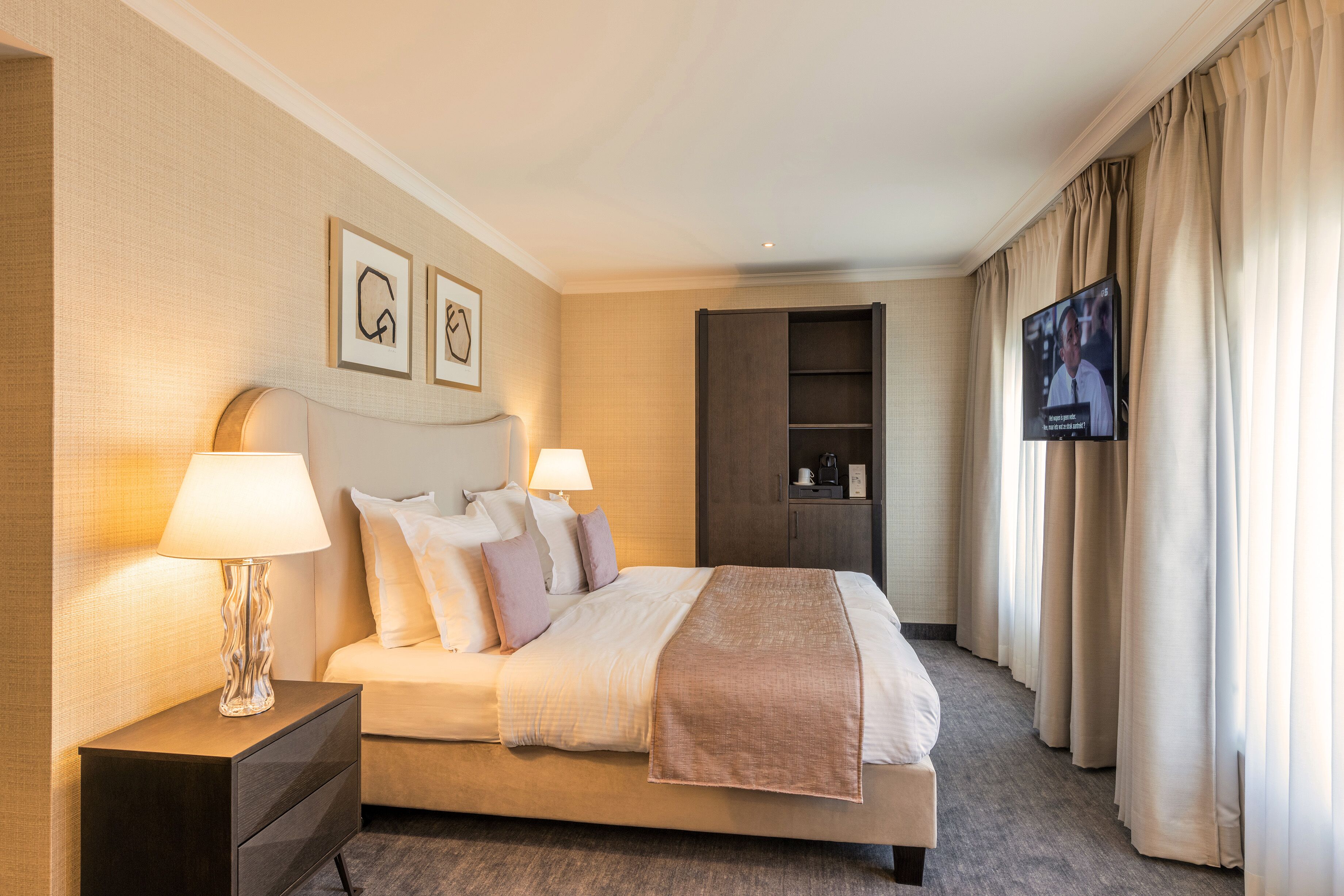 Una habitación de hotel con cama, dos lámparas de noche, televisión, armario y cuadros en la pared.