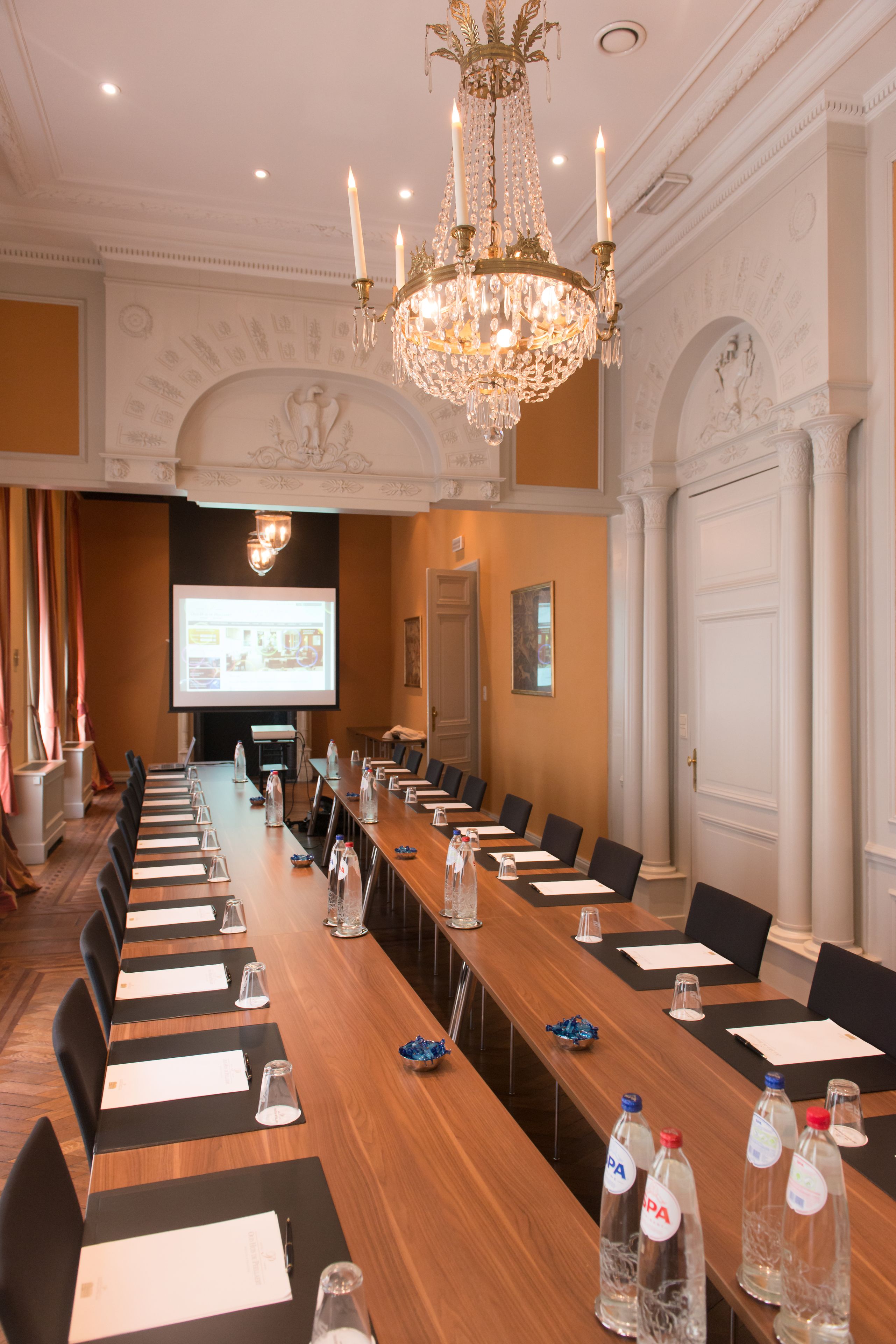 Un salón de conferencias con una larga mesa, sillas, una pantalla de proyector y una araña.