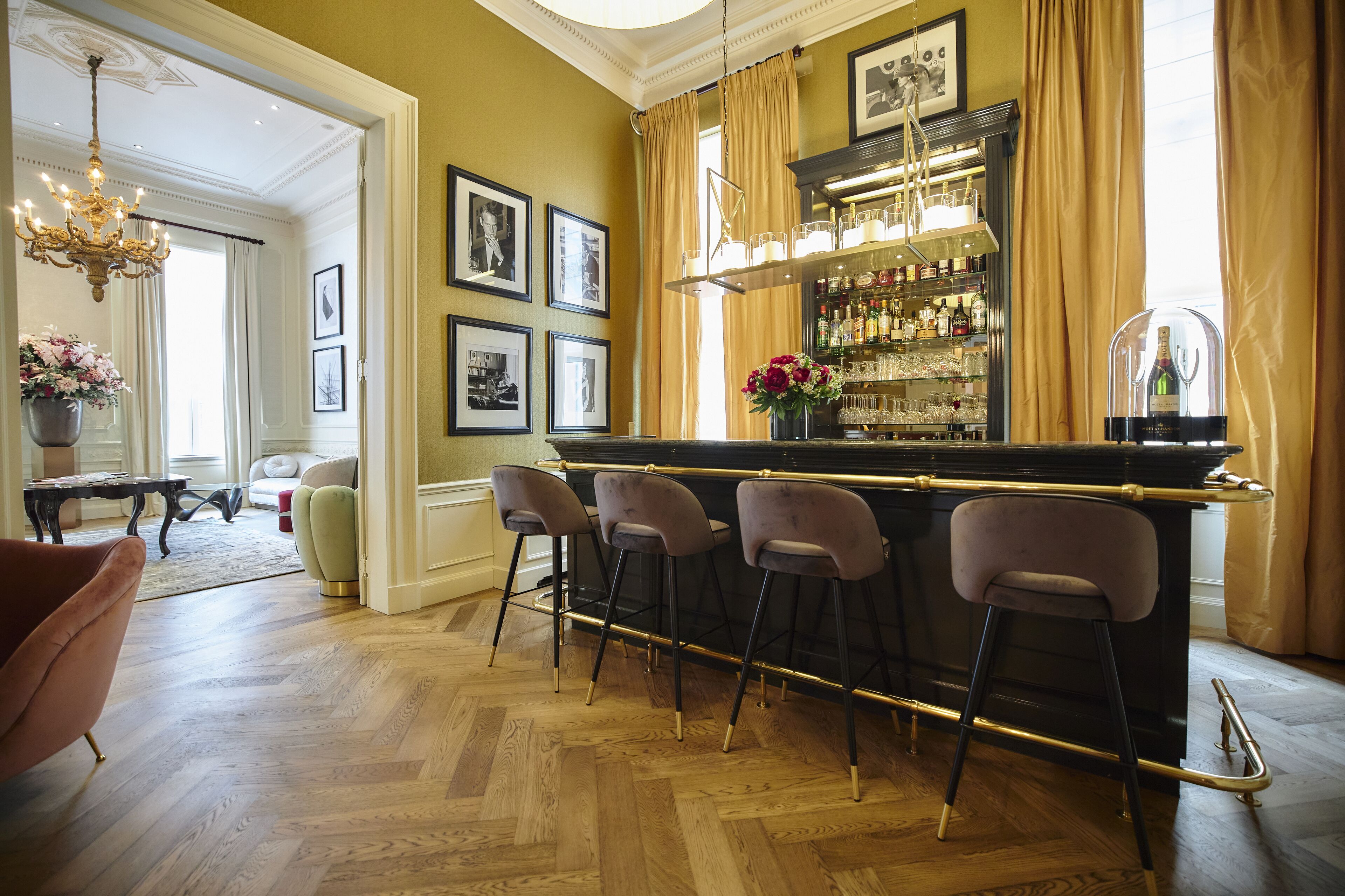A bar area with a wooden herringbone floor, bar stools, a wine glass display, and a chandelier.