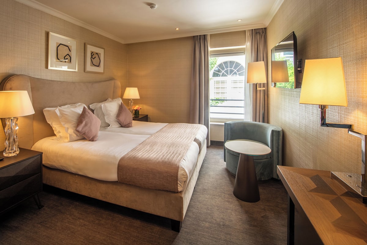 Ein Hotelzimmer mit einem großen Bett, zwei Nachttischen mit Lampen, einem kleinen runden Tisch, einem Sessel und einem Fenster mit Vorhängen.