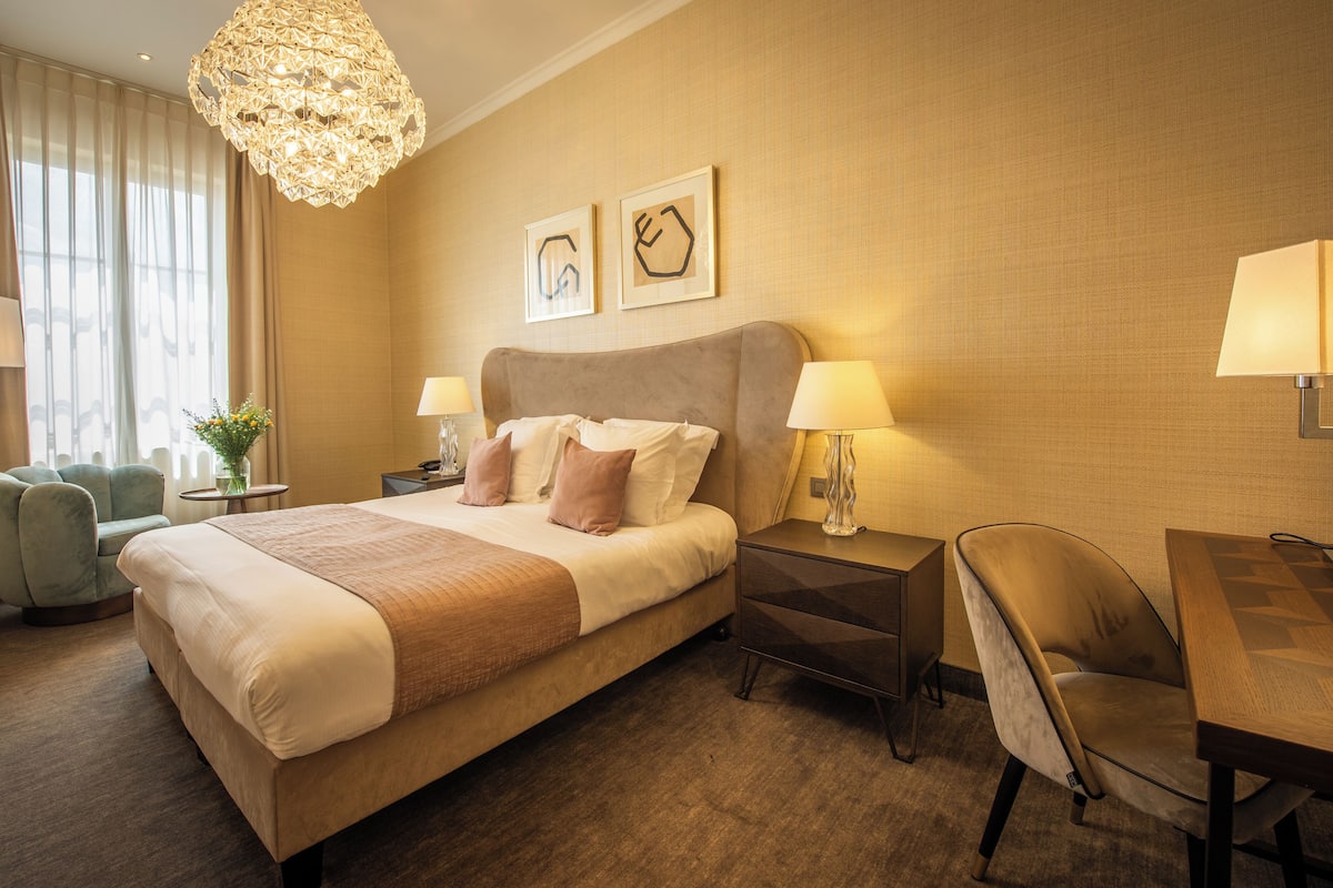 Ein Hotelzimmer mit einem großen Bett, Nachttischlampen, einem Schreibtisch und einem Sessel. An der Wand hängen zwei gerahmte Bilder und über dem Bett hängt ein Kronleuchter.