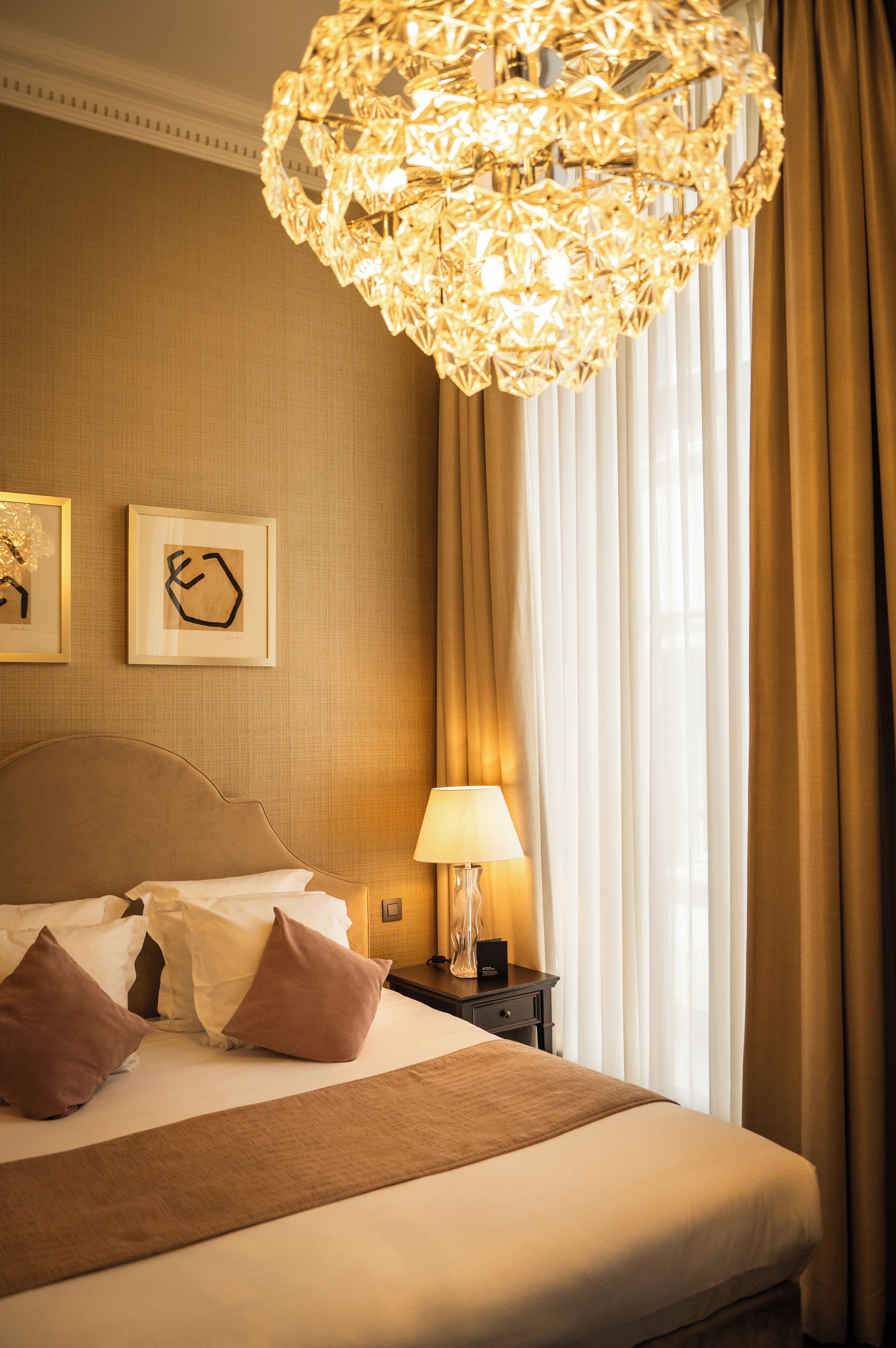 A hotel room with a large bed, a nightstand with a lamp, and a decorative chandelier.