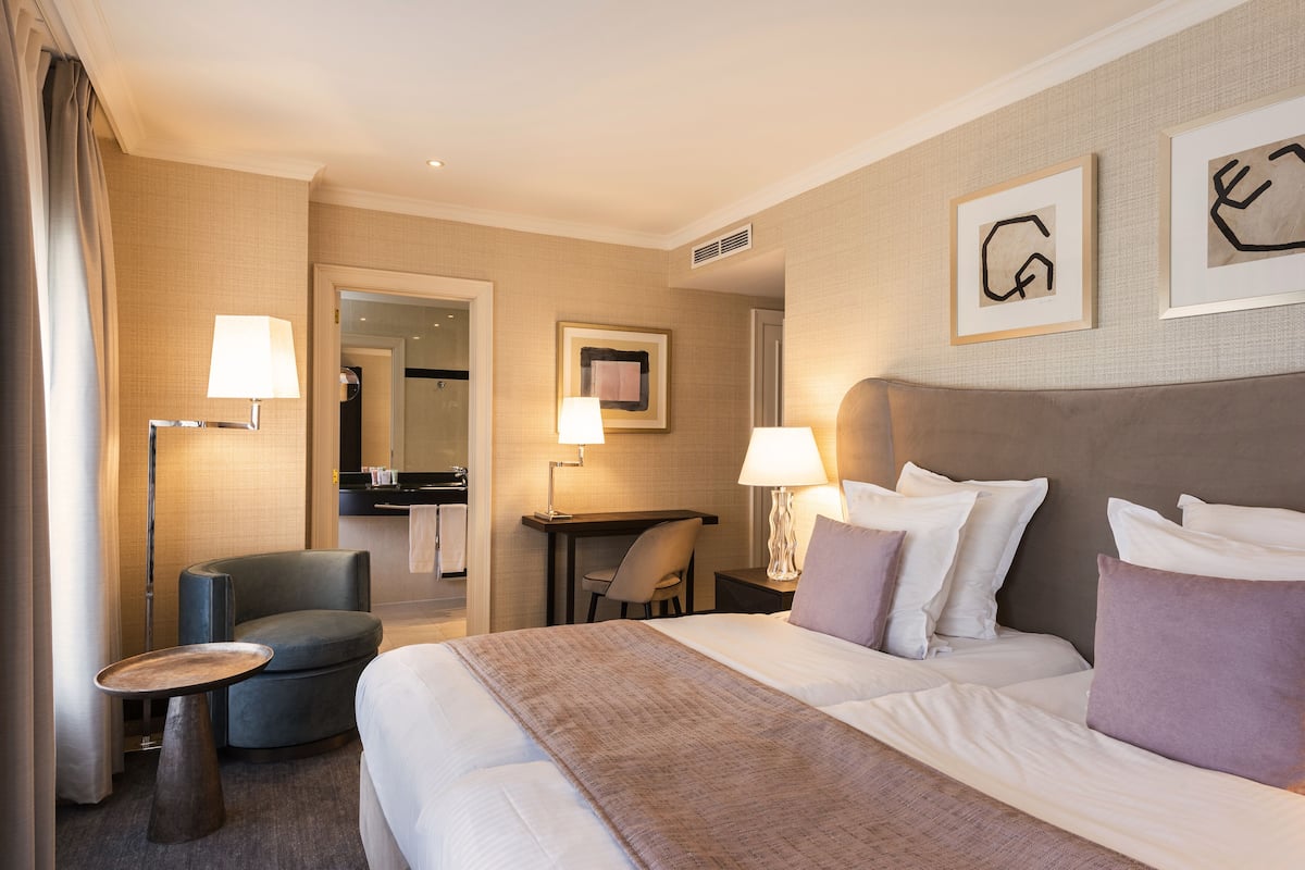 Ein Hotelzimmer mit einem großen Bett, zwei Nachttischen, einem Schreibtisch, einem Stuhl und einem kleinen runden Tisch. Auf den Nachttischen stehen jeweils Lampen und an der Wand hängt ein Bilderrahmen.