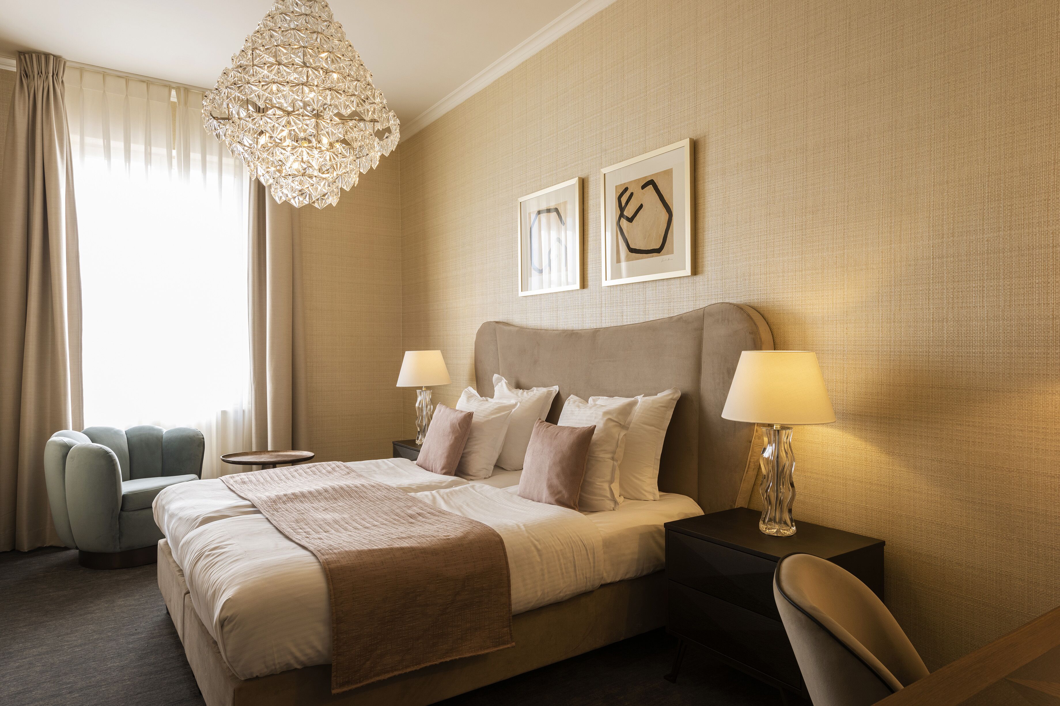 A hotel room with a large bed, two bedside tables with lamps, a decorative chandelier, and two framed pictures on the wall.