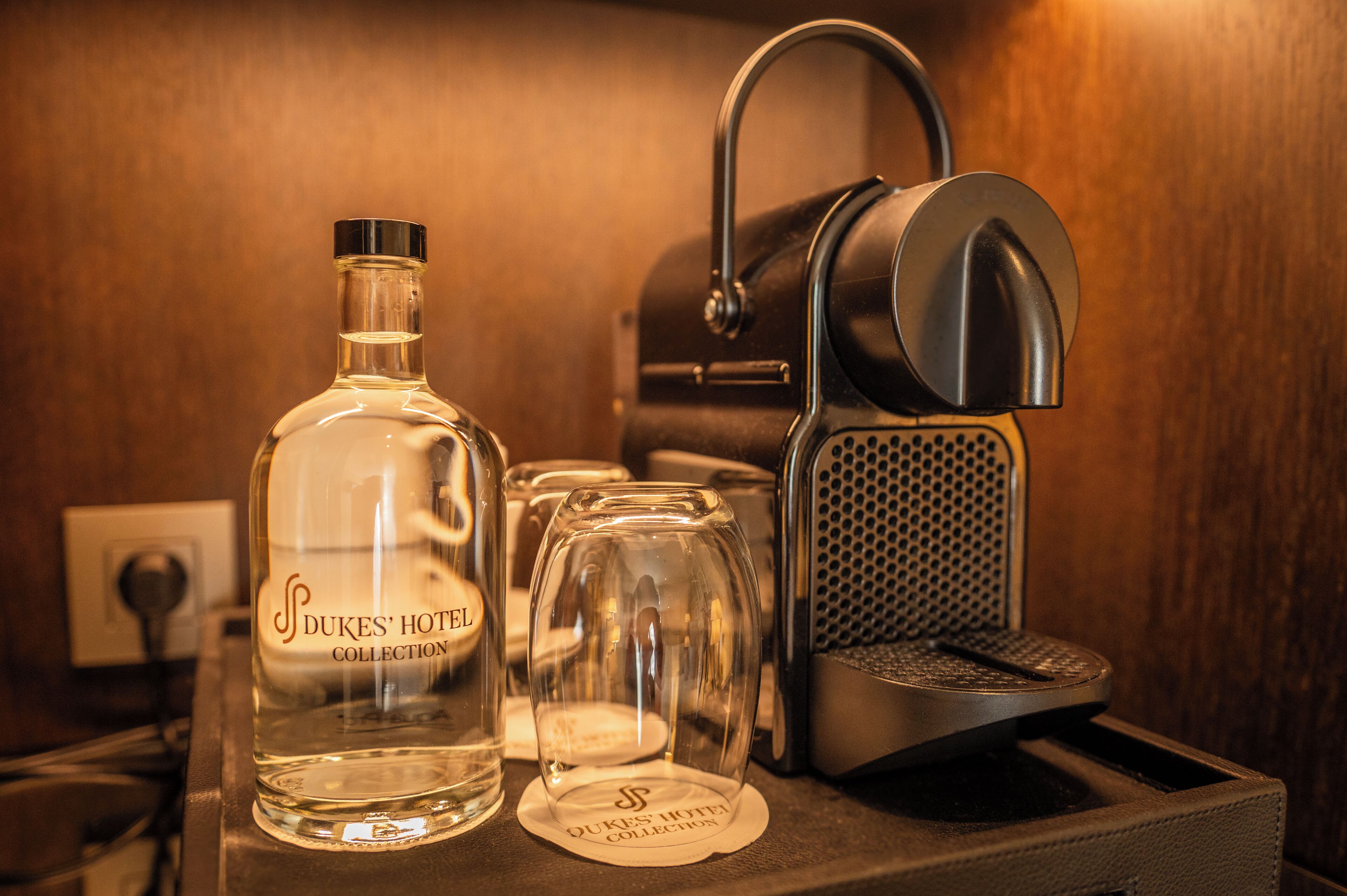 A bottle of Dukes' Hotel Collection, a coffee maker, and a glass on a countertop.