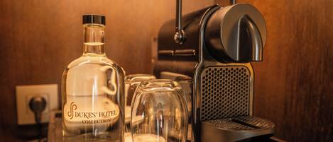 A bottle of Dukes' Hotel Collection, a coffee maker, and a glass on a countertop.
