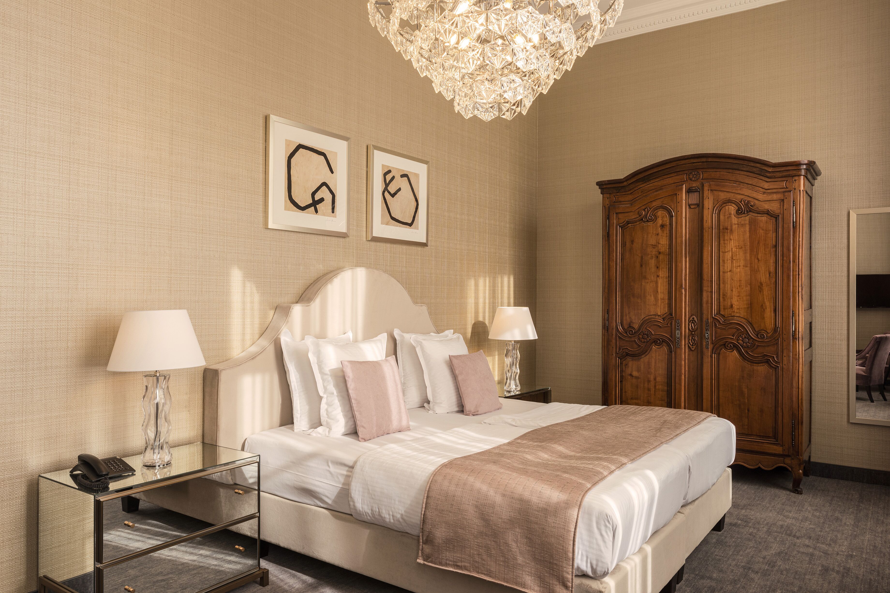 A hotel room with a large bed, bedside lamps, a wooden wardrobe, and two framed artworks on the wall.