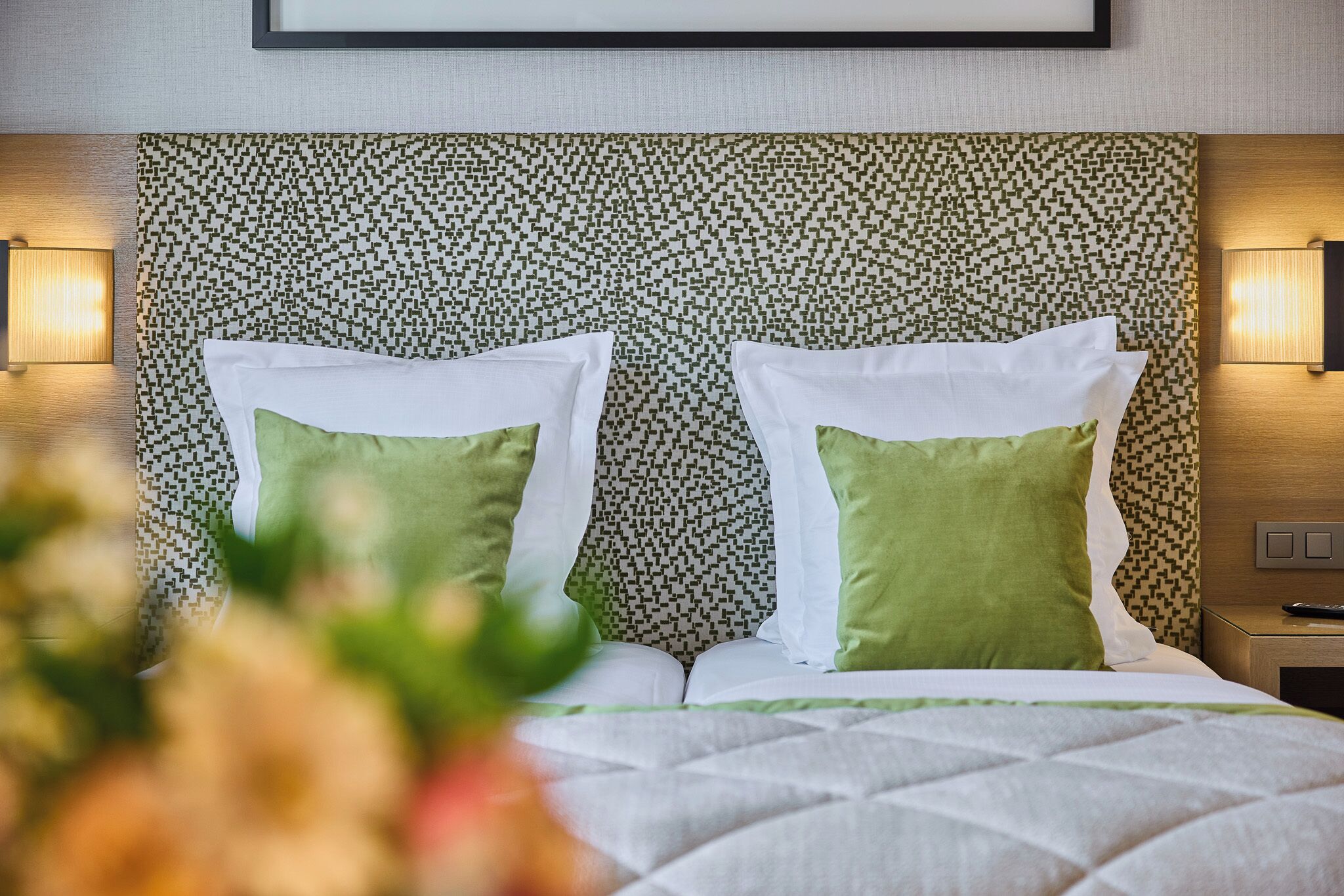 A hotel room with a patterned headboard, two pillows, a bed with a quilt, and wall-mounted lamps.