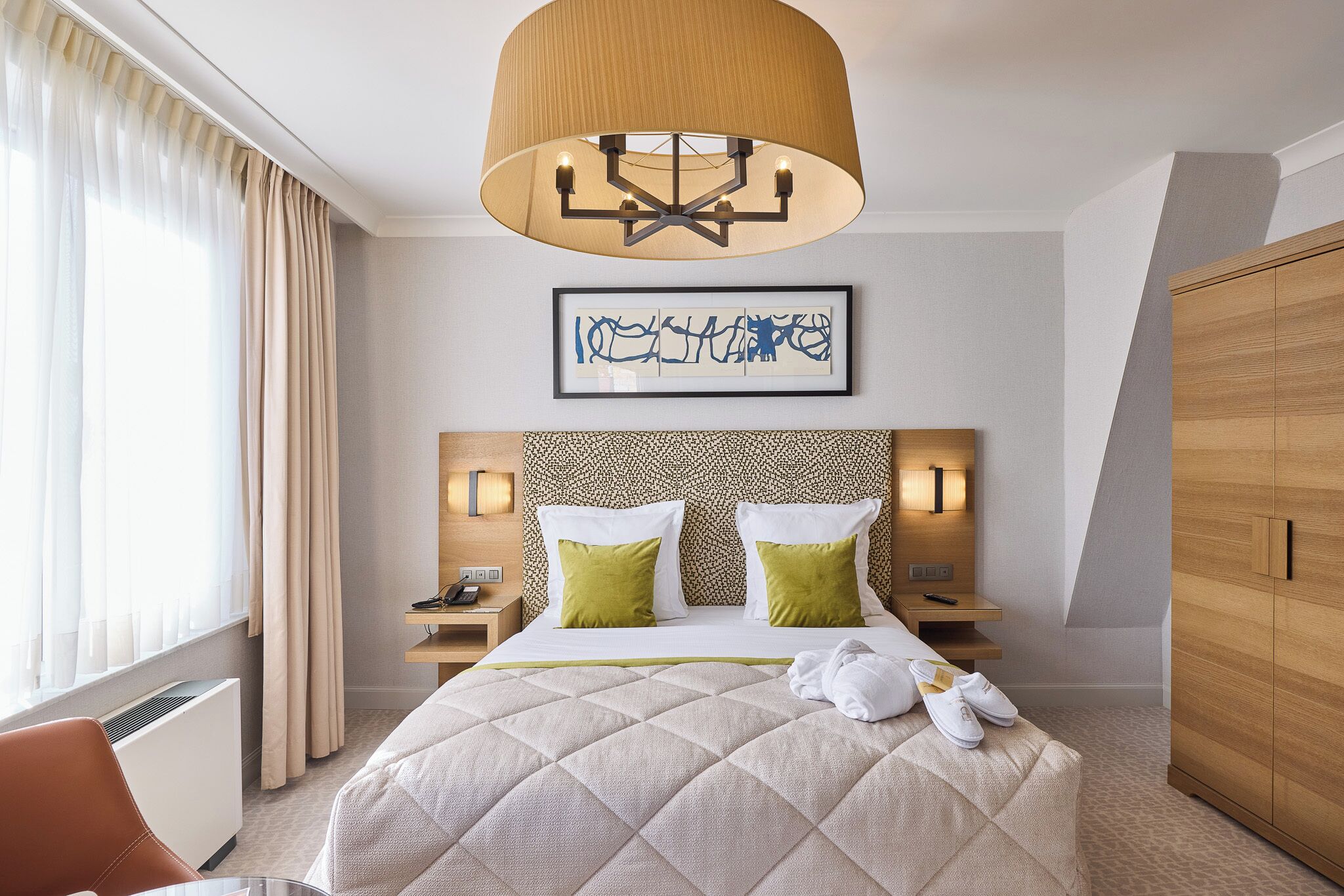 Une chambre d’hôtel avec un lit, un grand lustre, un tableau encadré, une armoire et une table de chevet avec un téléphone et une lampe.