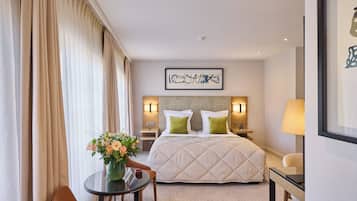 A hotel room with a large bed, a bedside table with a lamp, a chair, a small table with a vase of flowers, and a framed artwork on the wall.