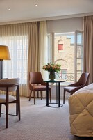 Habitación de hotel con cama, dos sillas, una mesita con un florero y vistas a un edificio desde una ventana abierta.